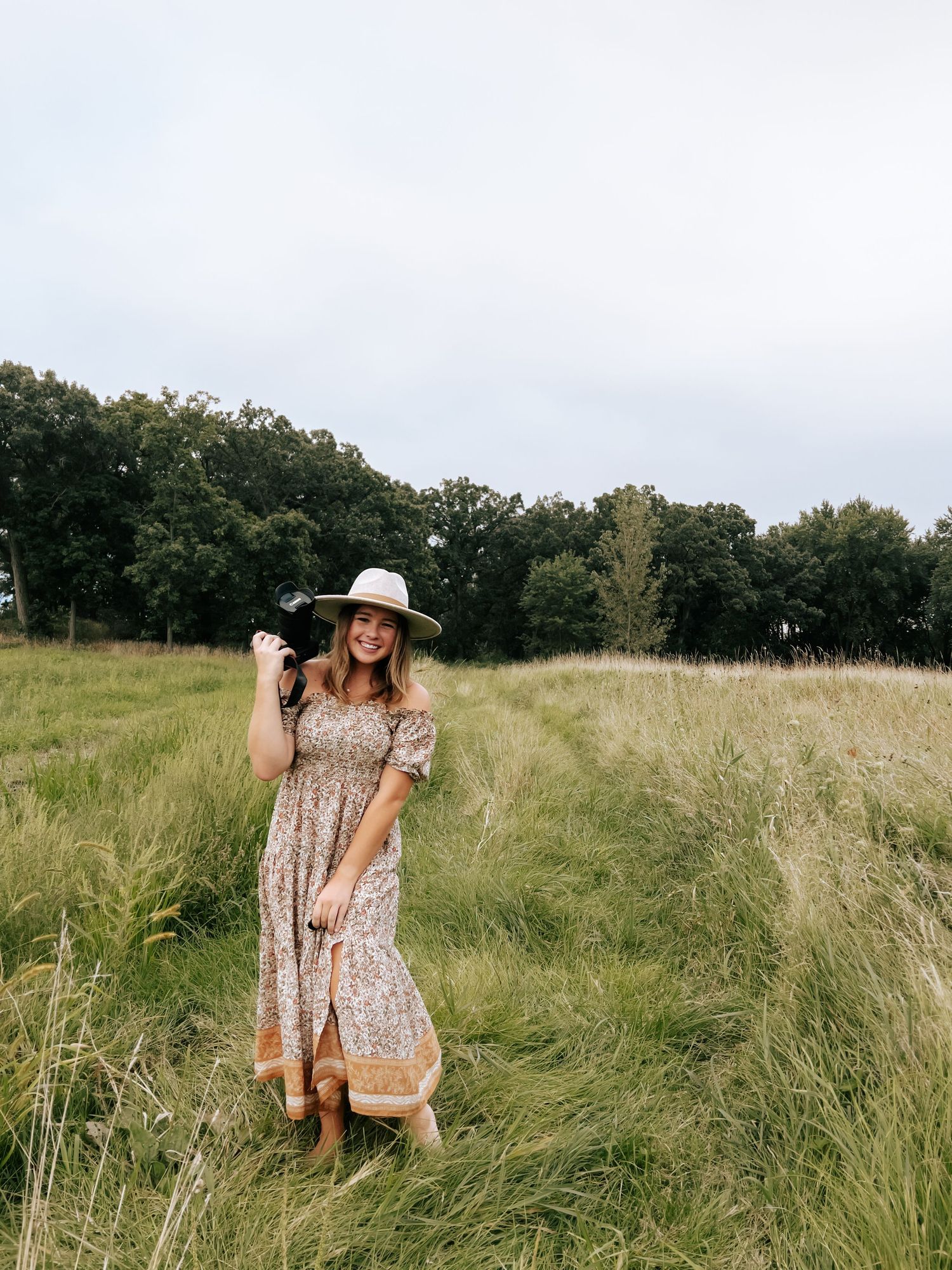 Woman in floral dress twirling in a sunlit meadow with tall grass and trees in background.