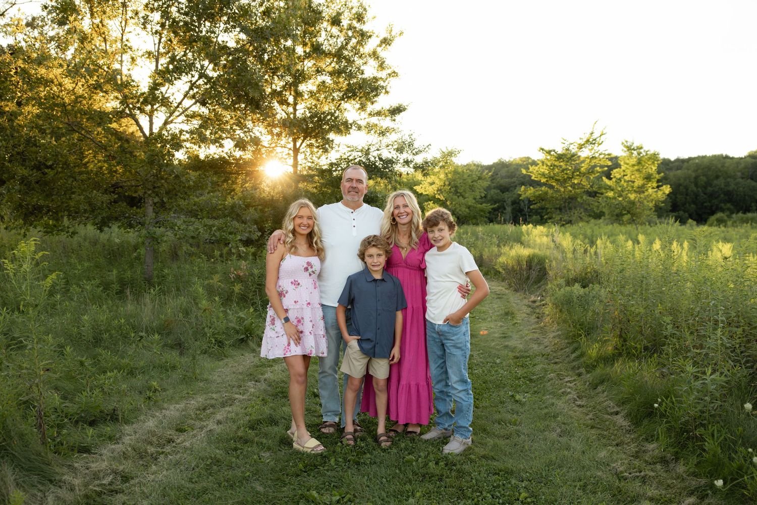 Family portrait in a grassy field with sunset light filtering through trees in background.