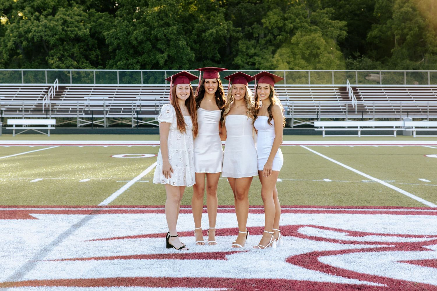 Four graduates in white dresses pose on a football field with bleachers behind them.