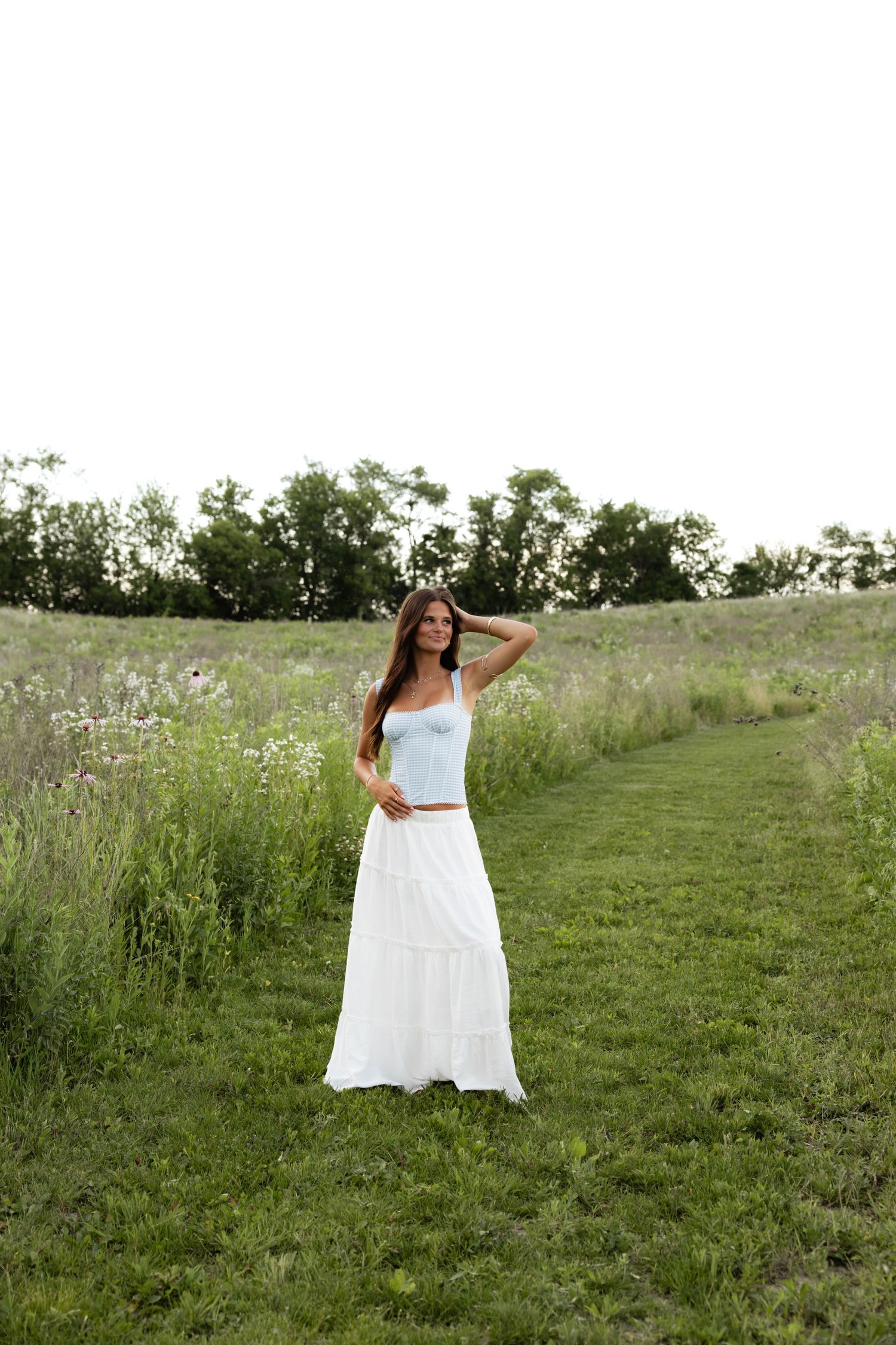 A woman in a white maxi dress stands in a meadow path surrounded by tall grass and wildflowers.