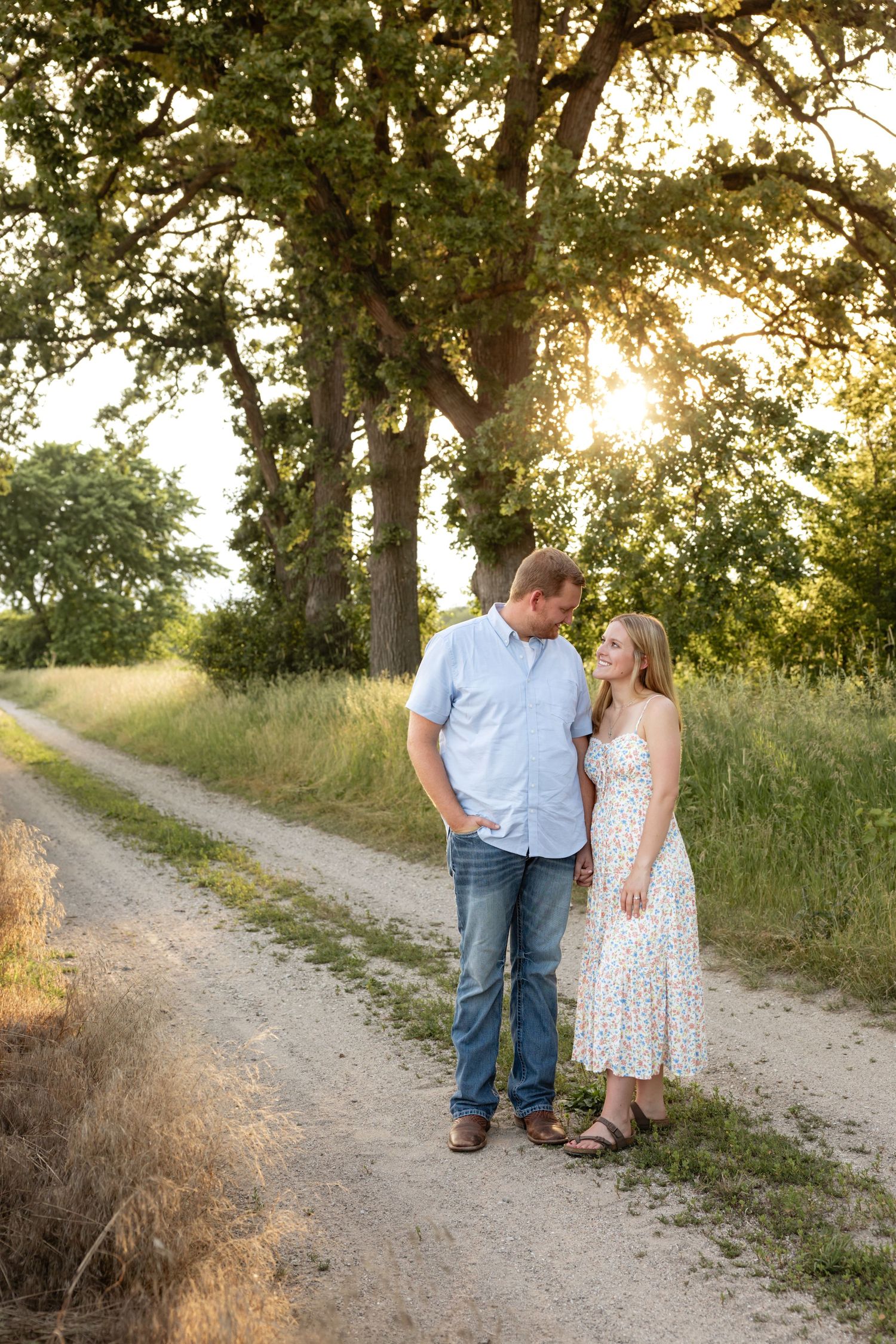 Couple walking together on rural dirt path with trees during sunset.
