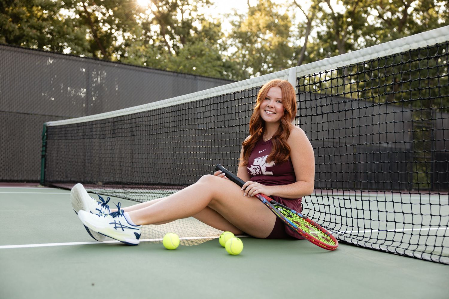 Person resting beside tennis court net with tennis equipment.