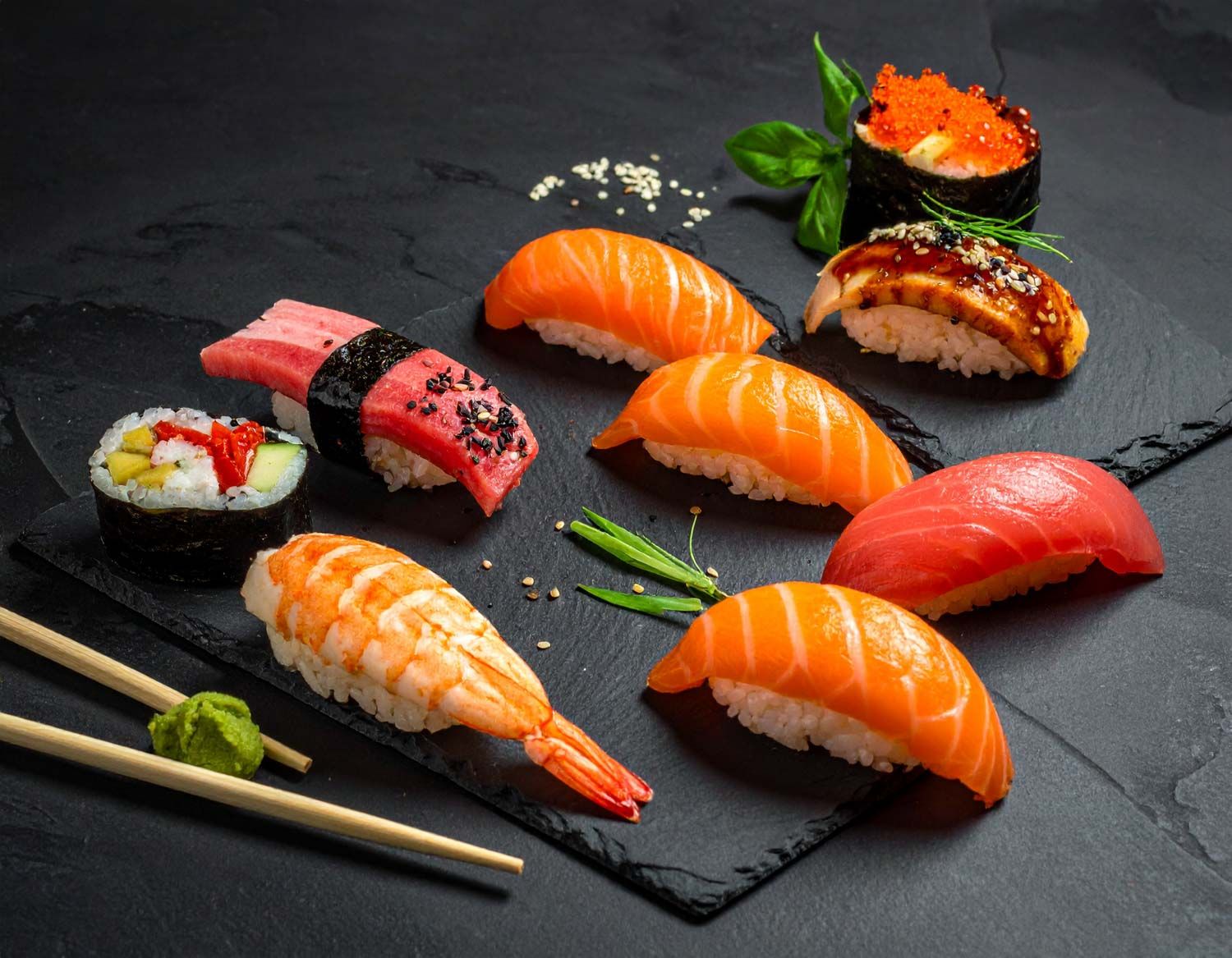 Pezzi assortiti di sushi fresco e nigiri disposti su ardesia nera con bacchette.