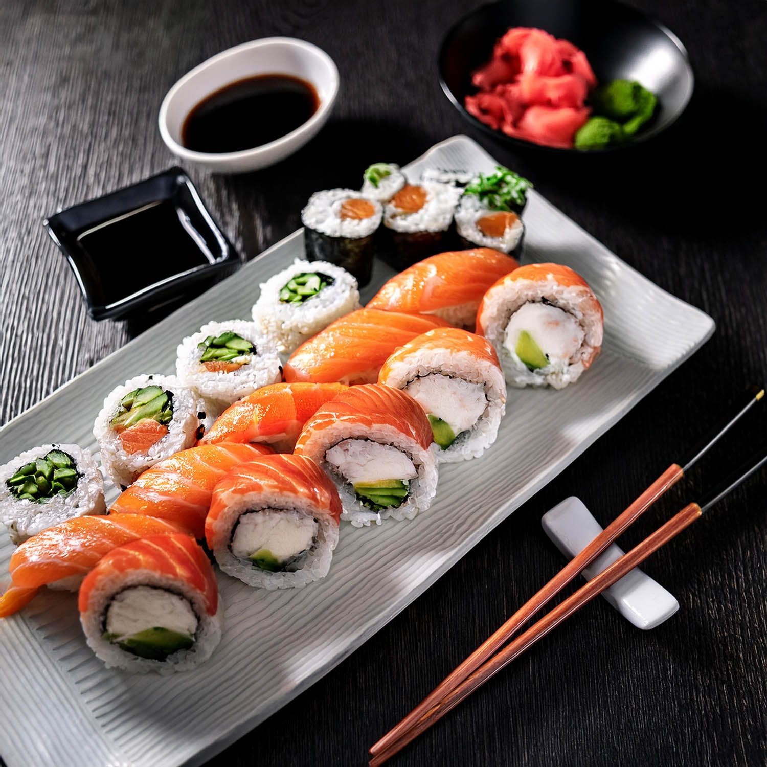 Roll freschi e nigiri disposti su un lungo piatto bianco con bacchette e salsa di soia.