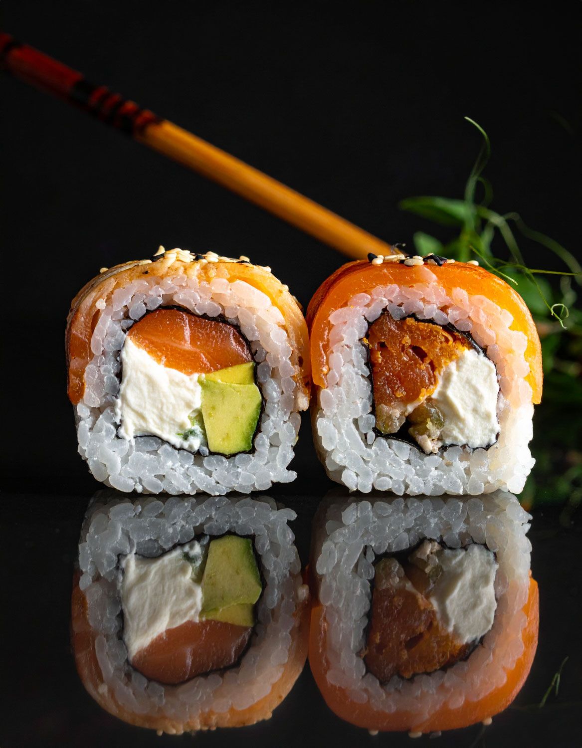 Primo piano di due pezzi di California roll con salmone e avocado riflessi su una superficie nera.