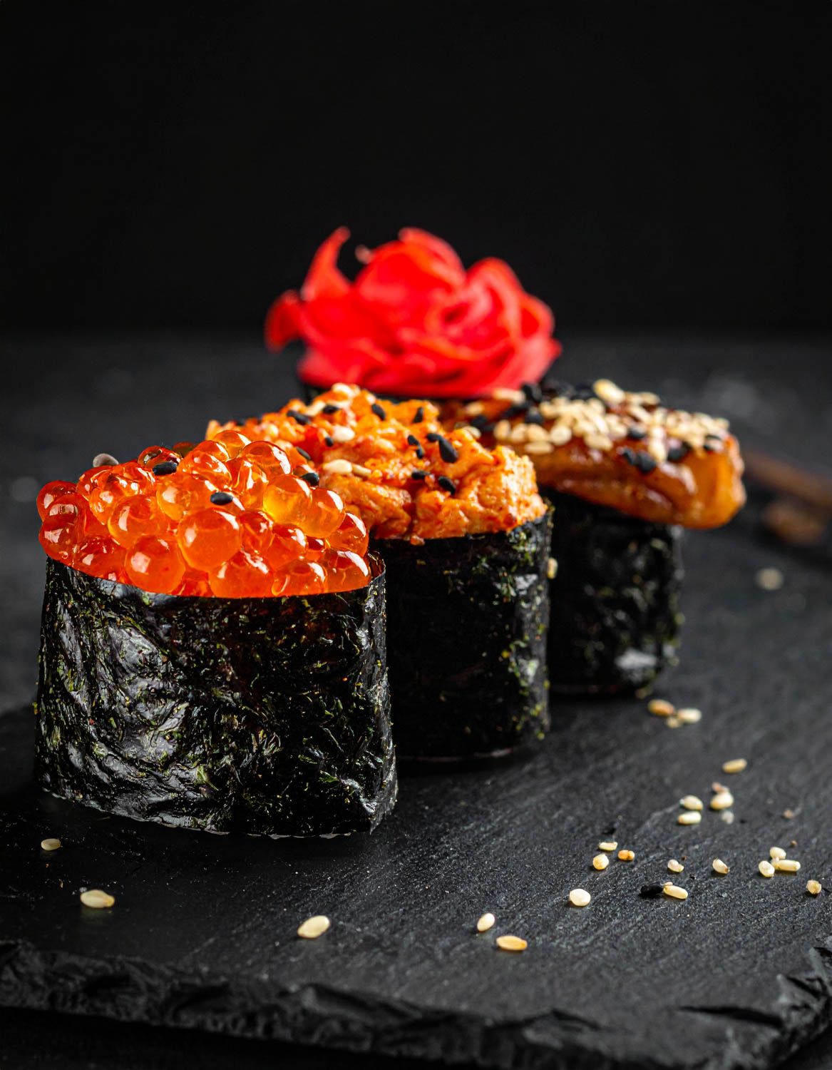 Tre roll di sushi gunkan guarniti con uova di salmone e zenzero rosso su ardesia nera.