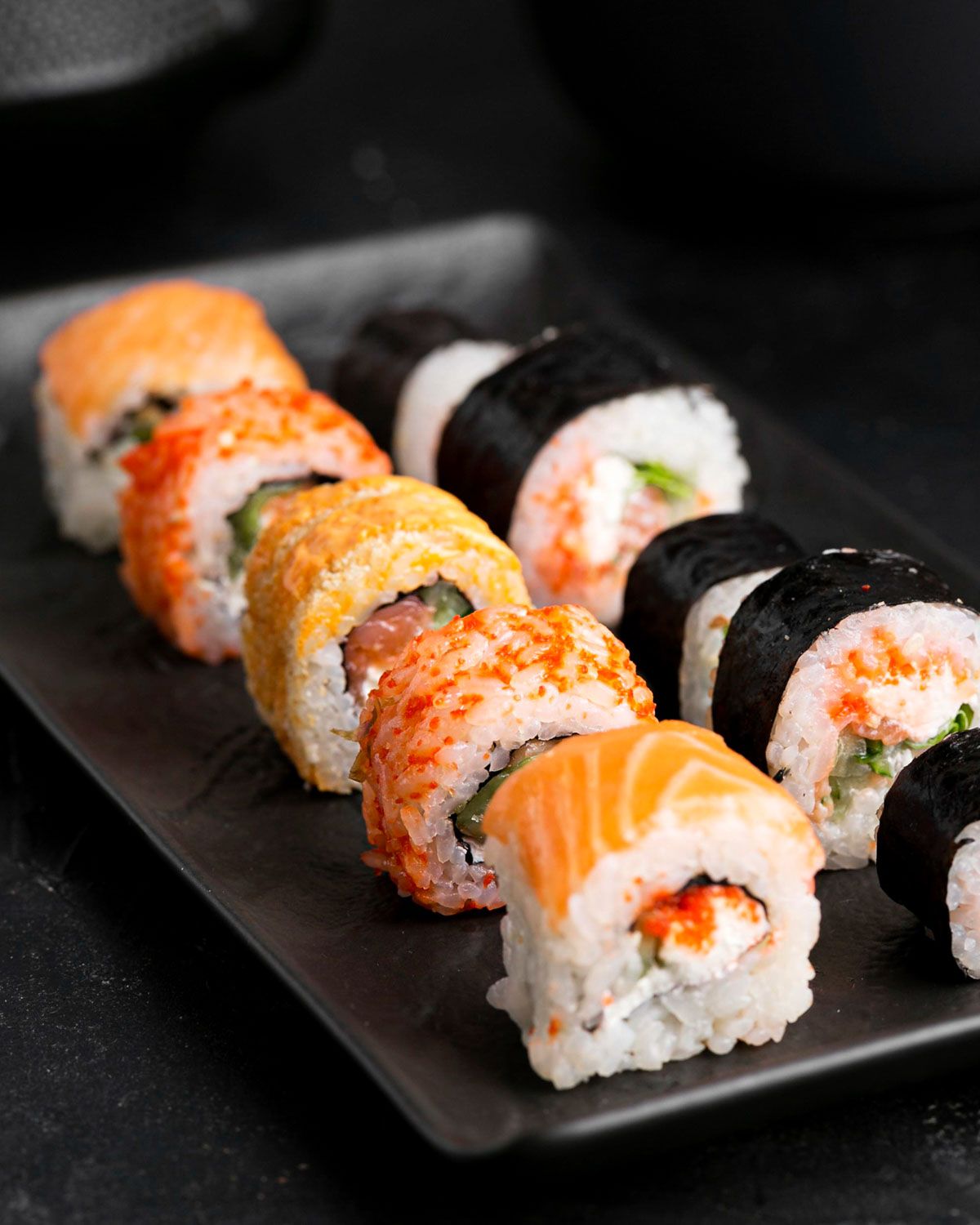 Assortimento colorato di roll con salmone, riso e alga nori su piatto nero.