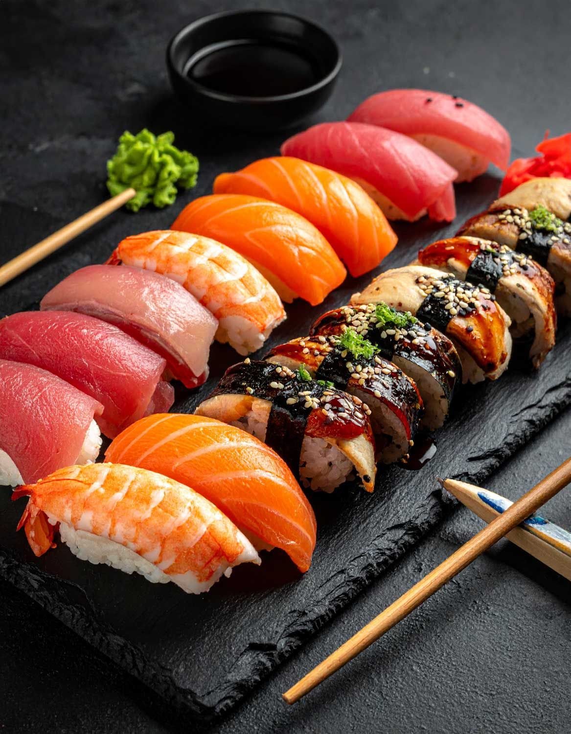 Assortimento colorato di nigiri freschi con salmone, tonno e guarnizioni disposti su piatti di ardesia nera.