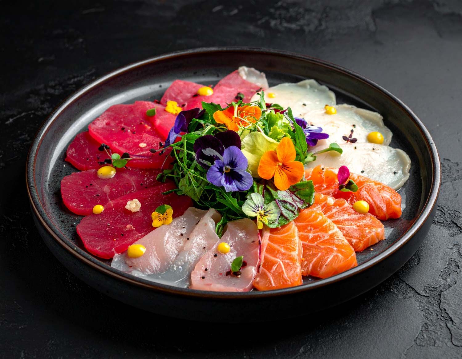 Presentazione artistica di sashimi con fiori eduli e micro-ortaggi su piatti neri.