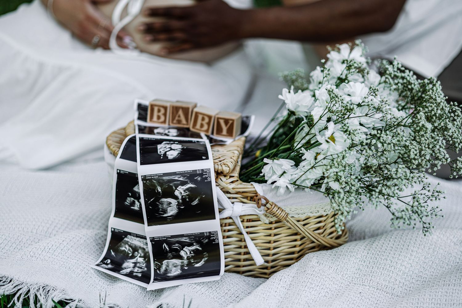 Ultrasound photos and baby wooden blocks beside a flower basket at a maternity photoshoot on a white blanket.