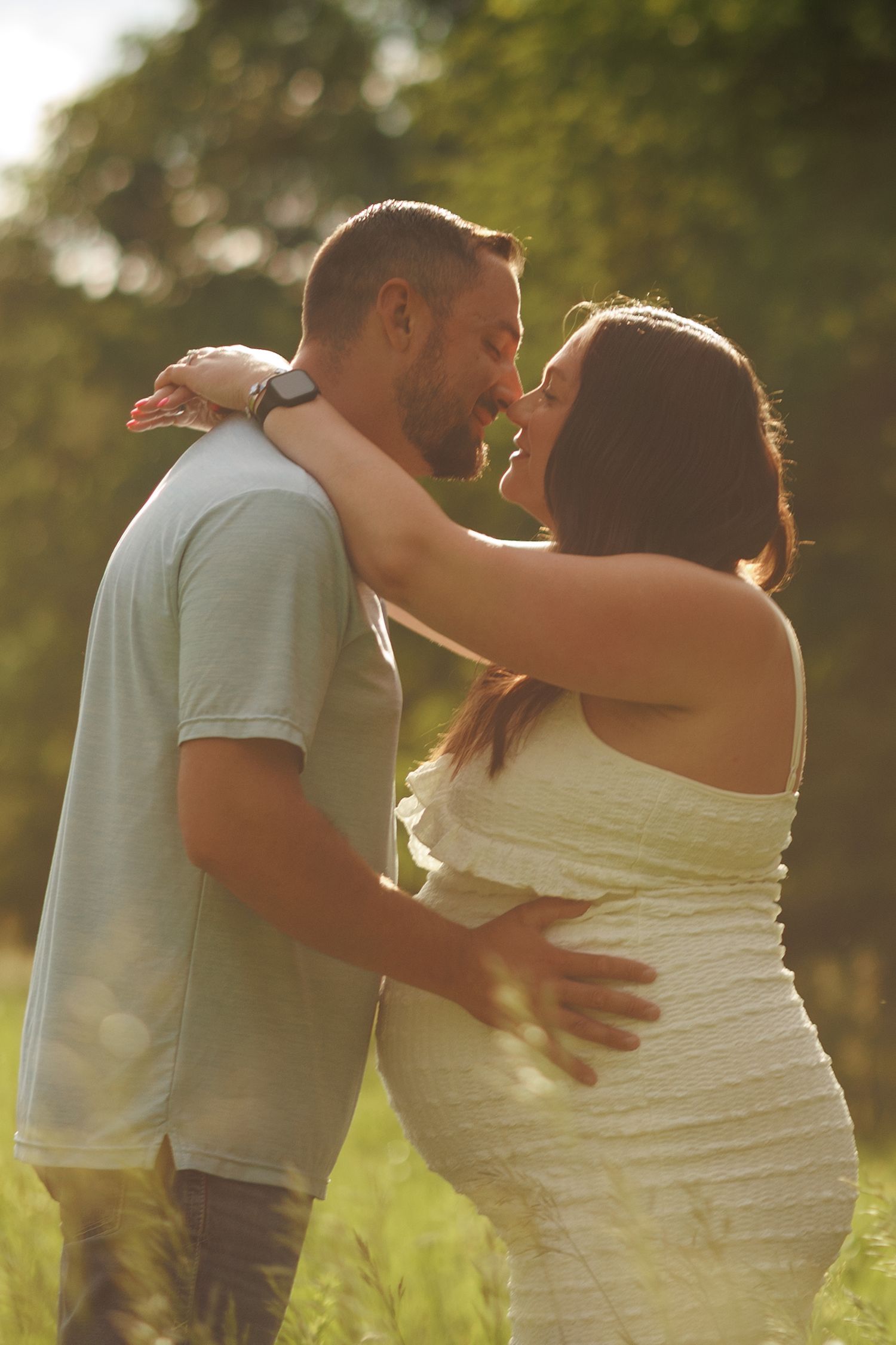 Couple sharing a tender moment outdoors, pregnant woman in white dress embraced by partner in sunlit green field.