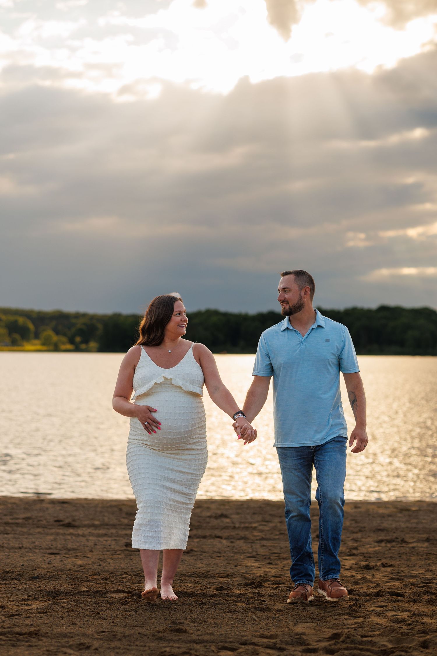 Pregnant couple holding hands on sandy beach at sunset, woman in white dress touching baby bump, man in blue shirt and jeans.