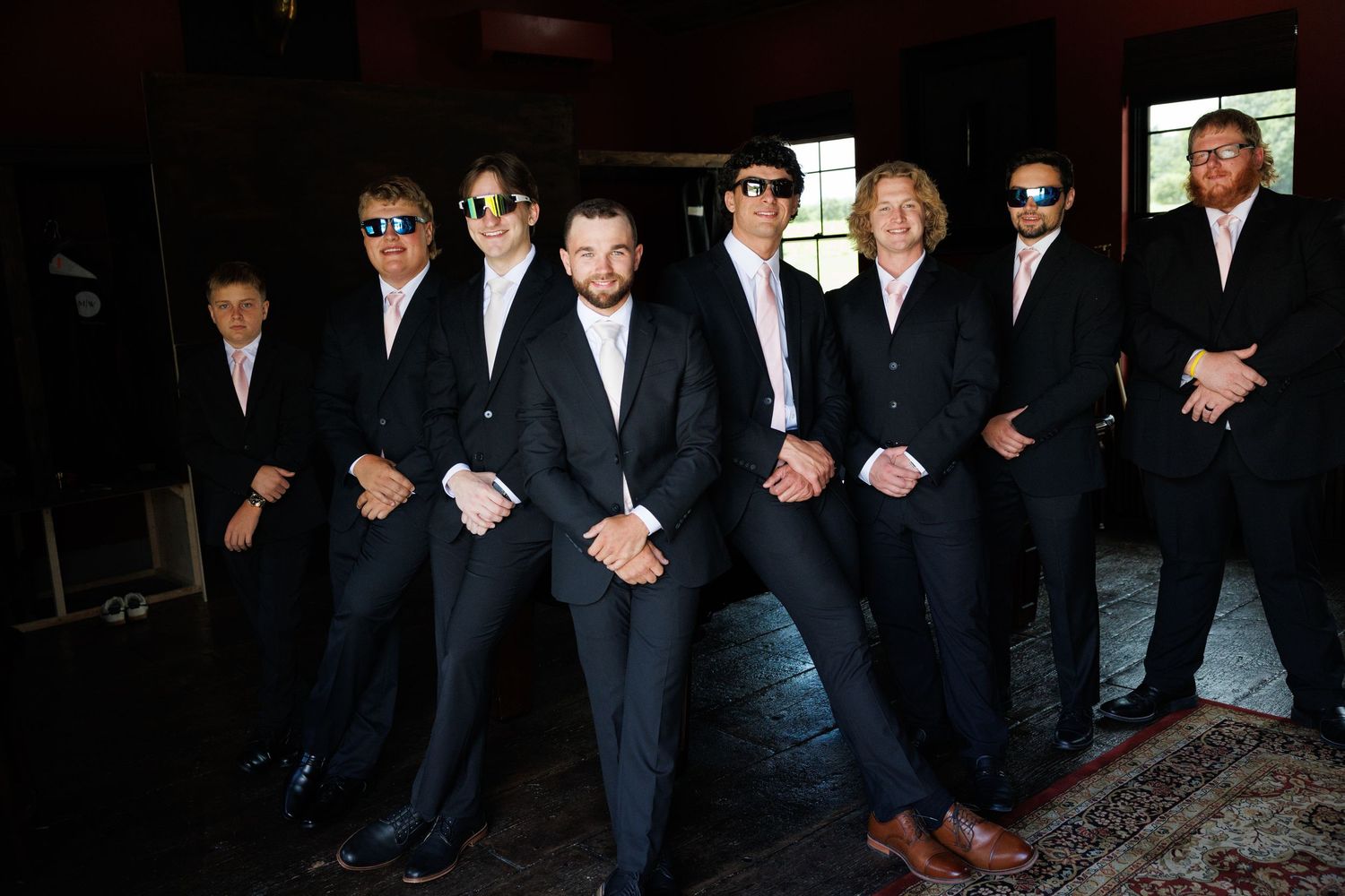 Group of eight men in matching dark suits and sunglasses posing confidently indoors for a formal occasion.