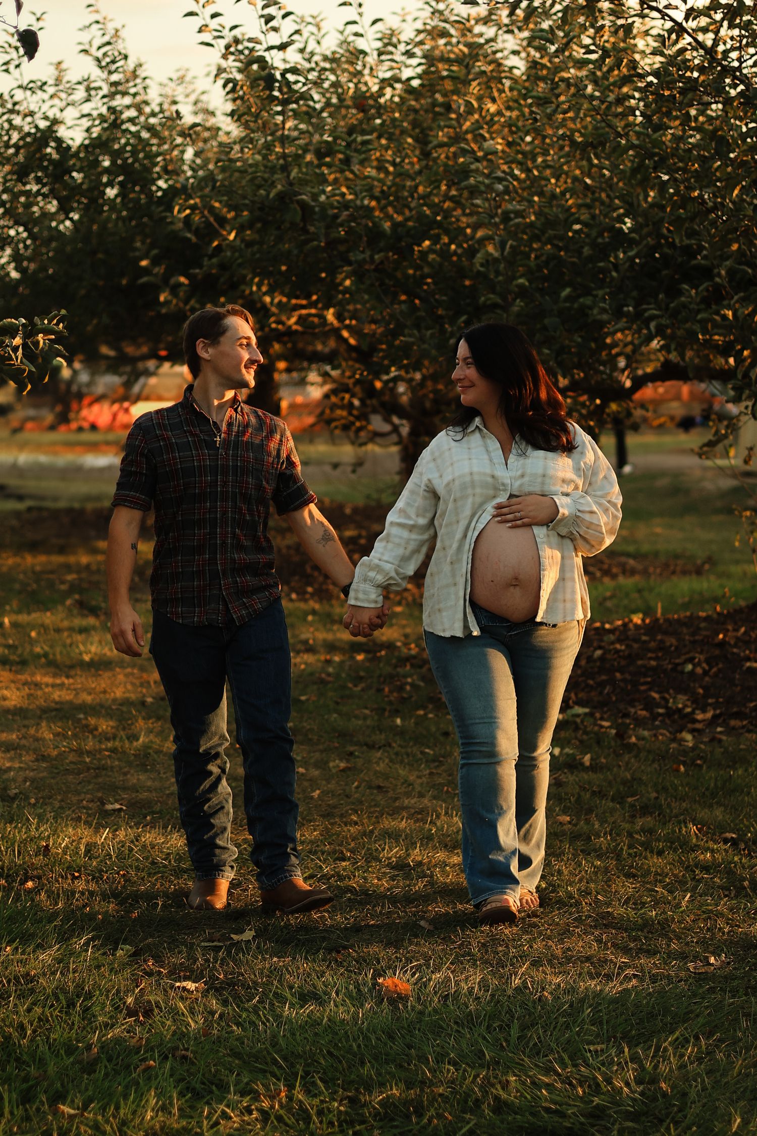 Expecting couple holding hands and laughing in an orchard at sunset, woman cradling her baby bump in jeans.