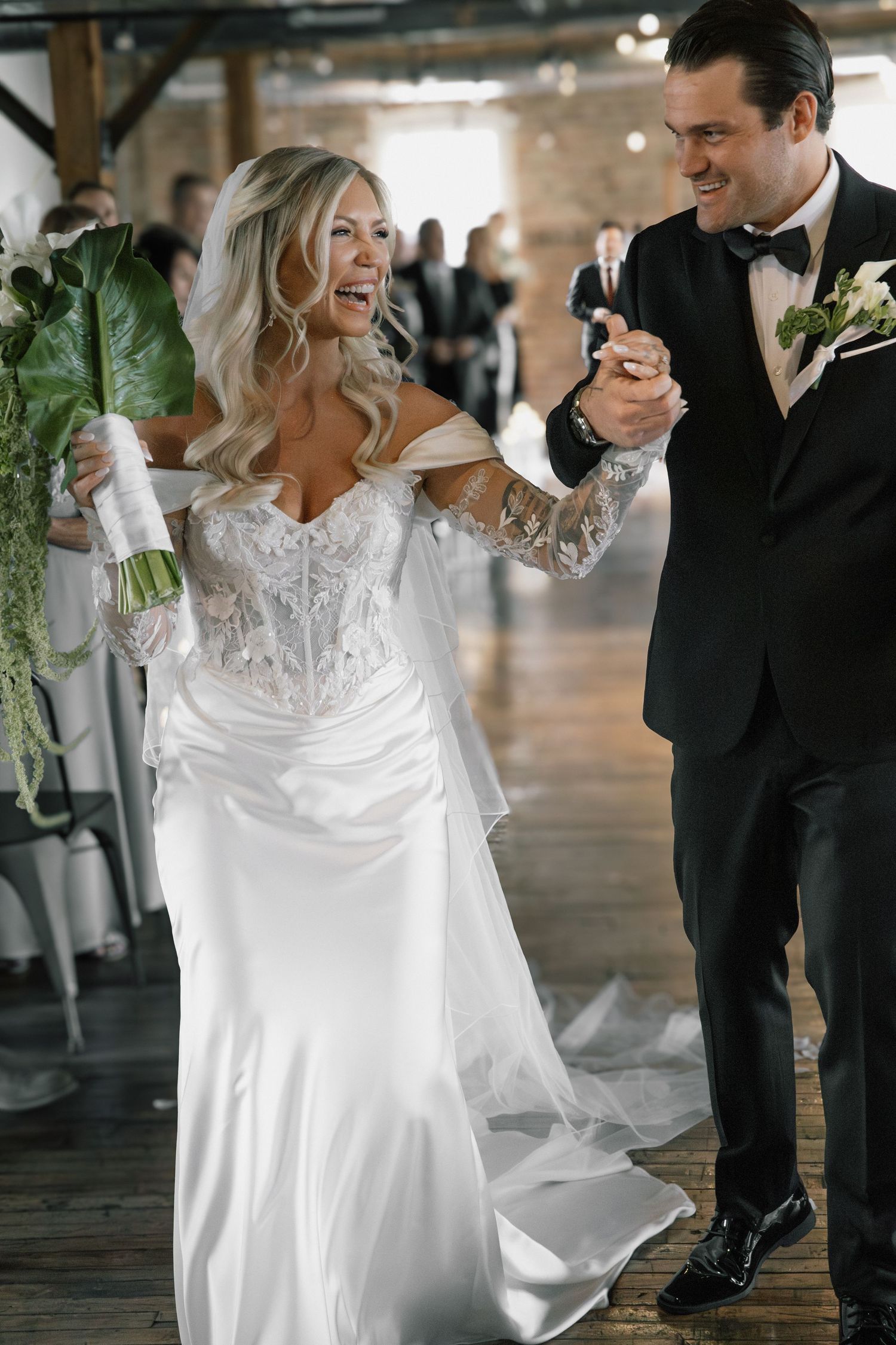 Smiling bride and groom walk hand in hand down aisle in rustic brick loft wedding venue.