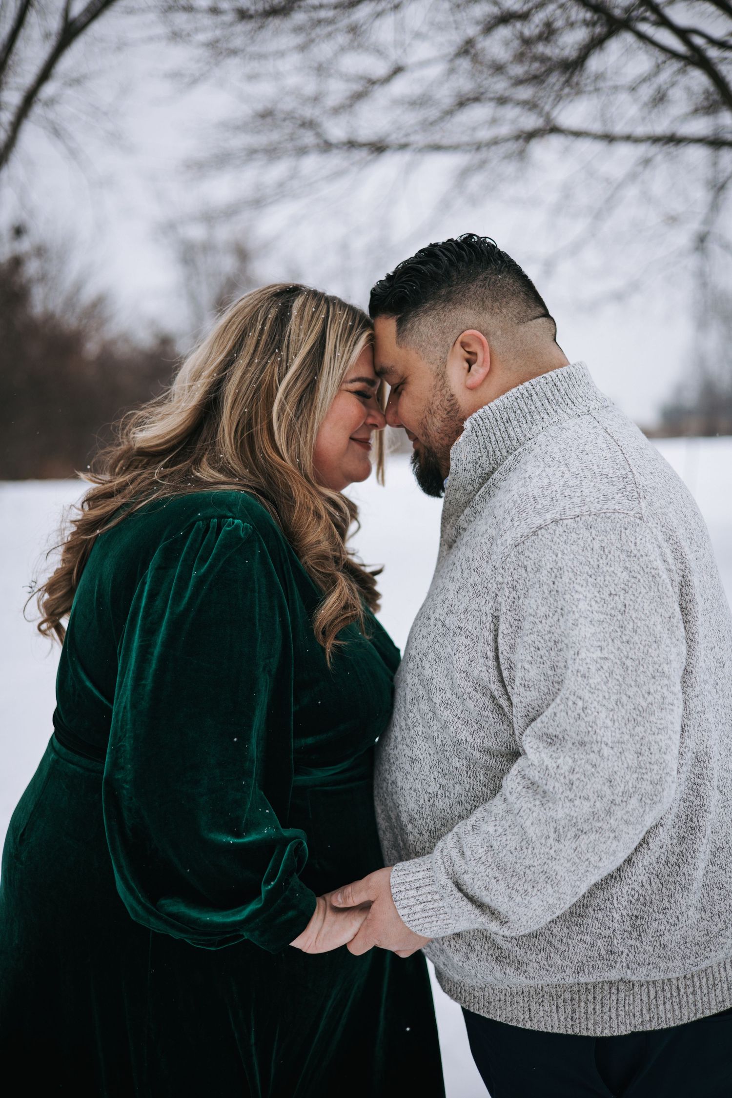 Expecting couple touching foreheads tenderly in snowy winter setting, woman in green velvet dress.