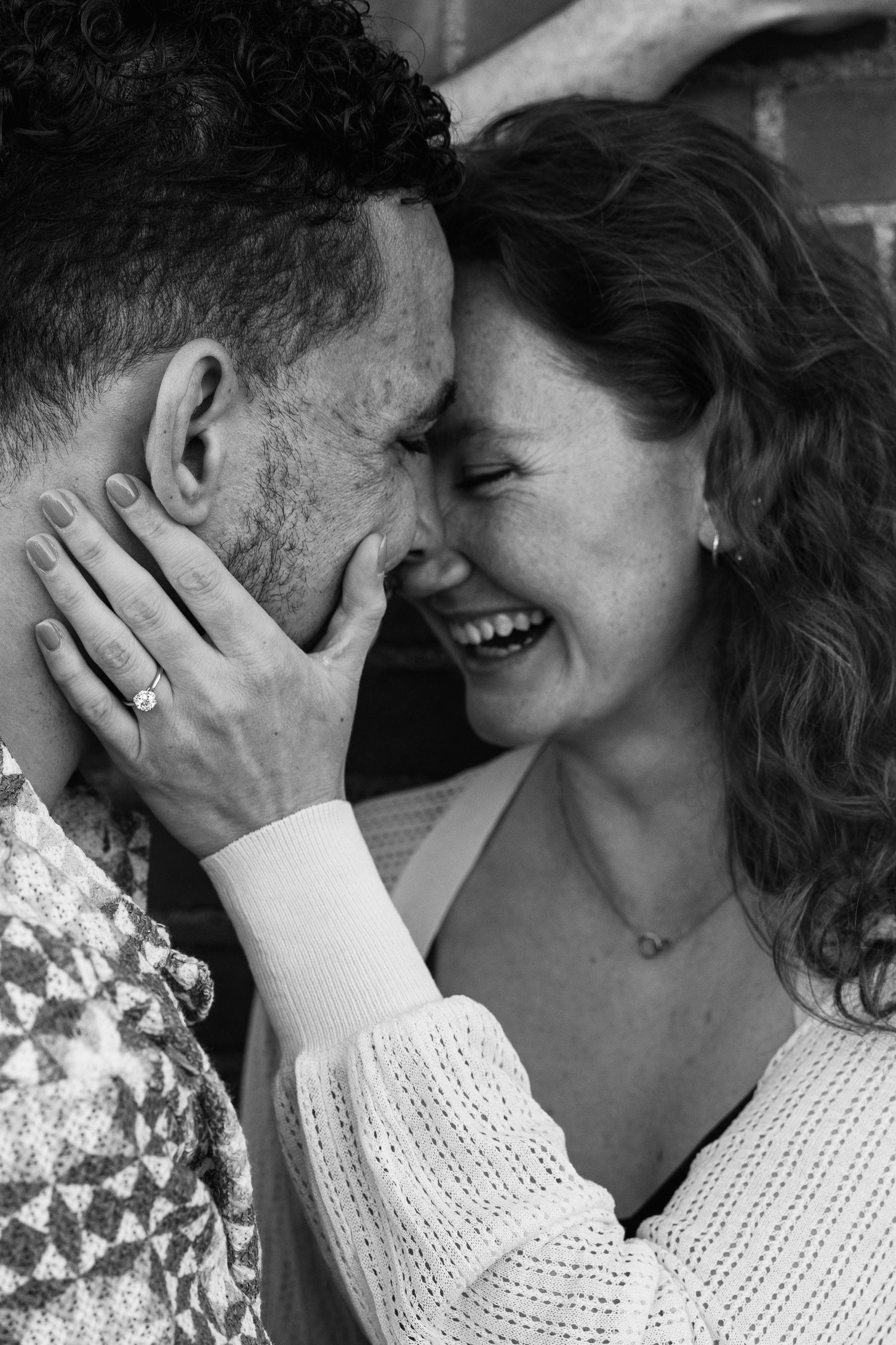 Black and white close-up of a smiling woman with freckles touching a mans face tenderly, wearing an engagement ring.
