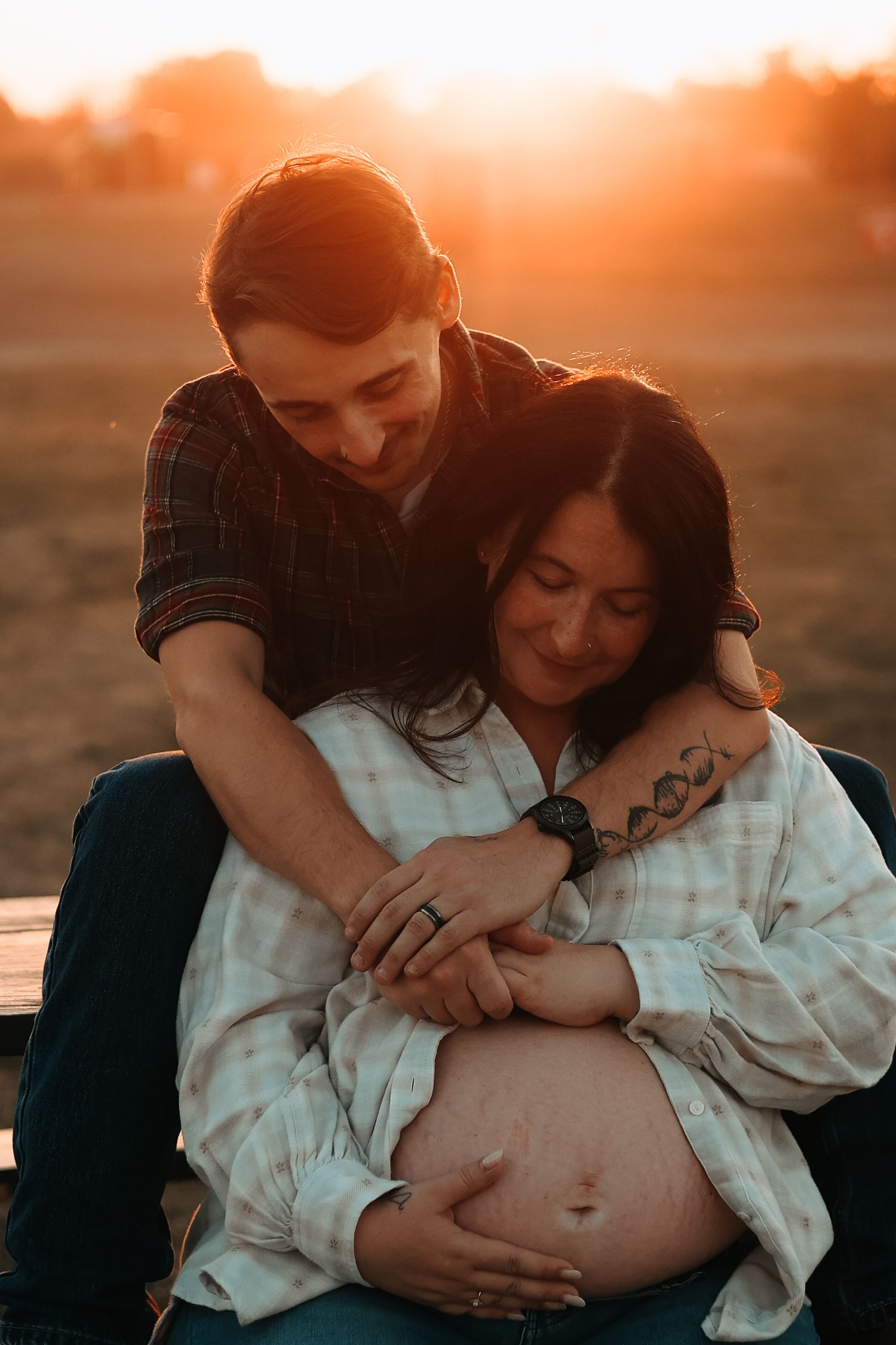 A couple embraces and holds a pregnant belly while sitting outdoors during a warm golden sunset.