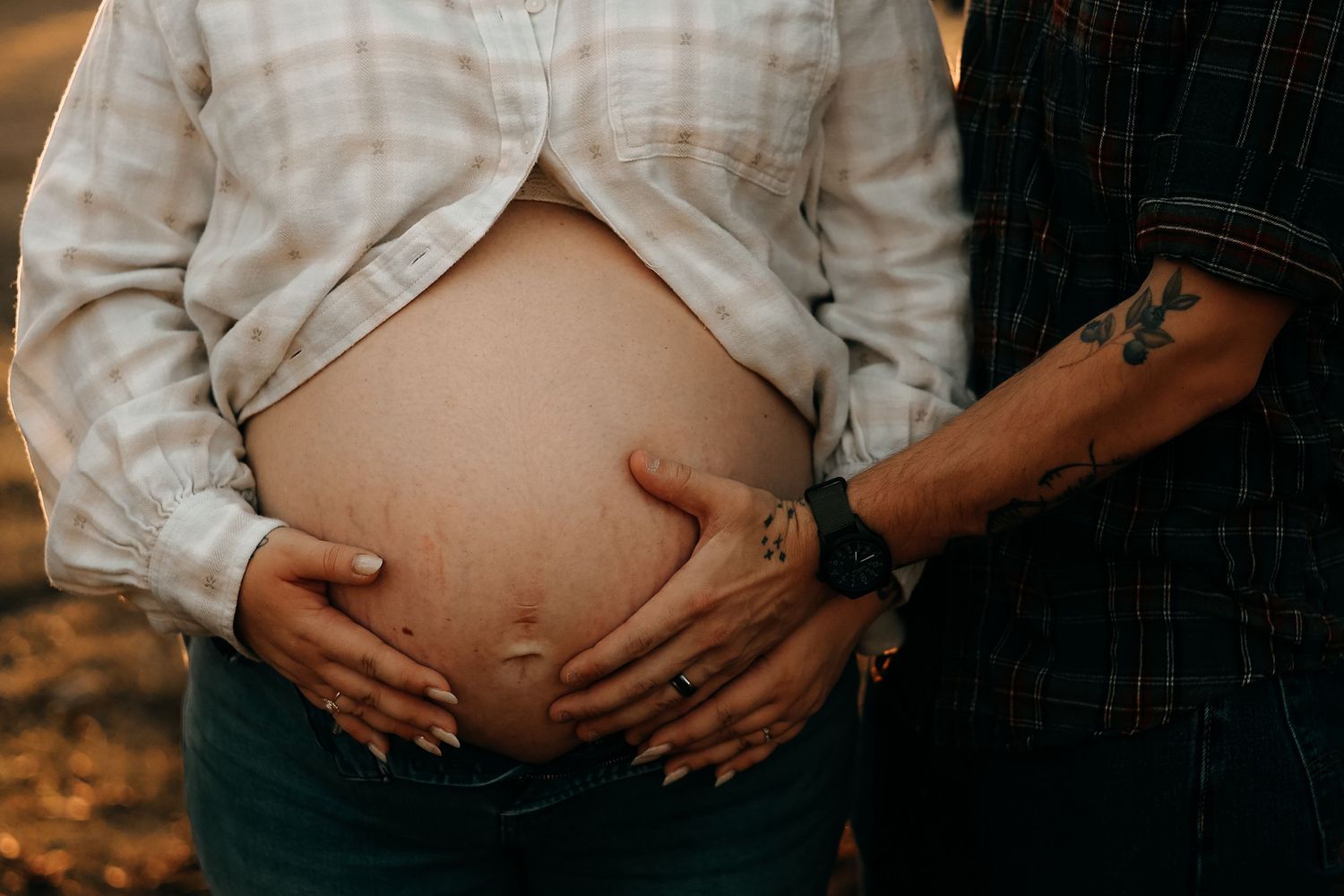 Close-up of pregnant belly with tattooed partner placing hand alongside mothers hands at golden hour.