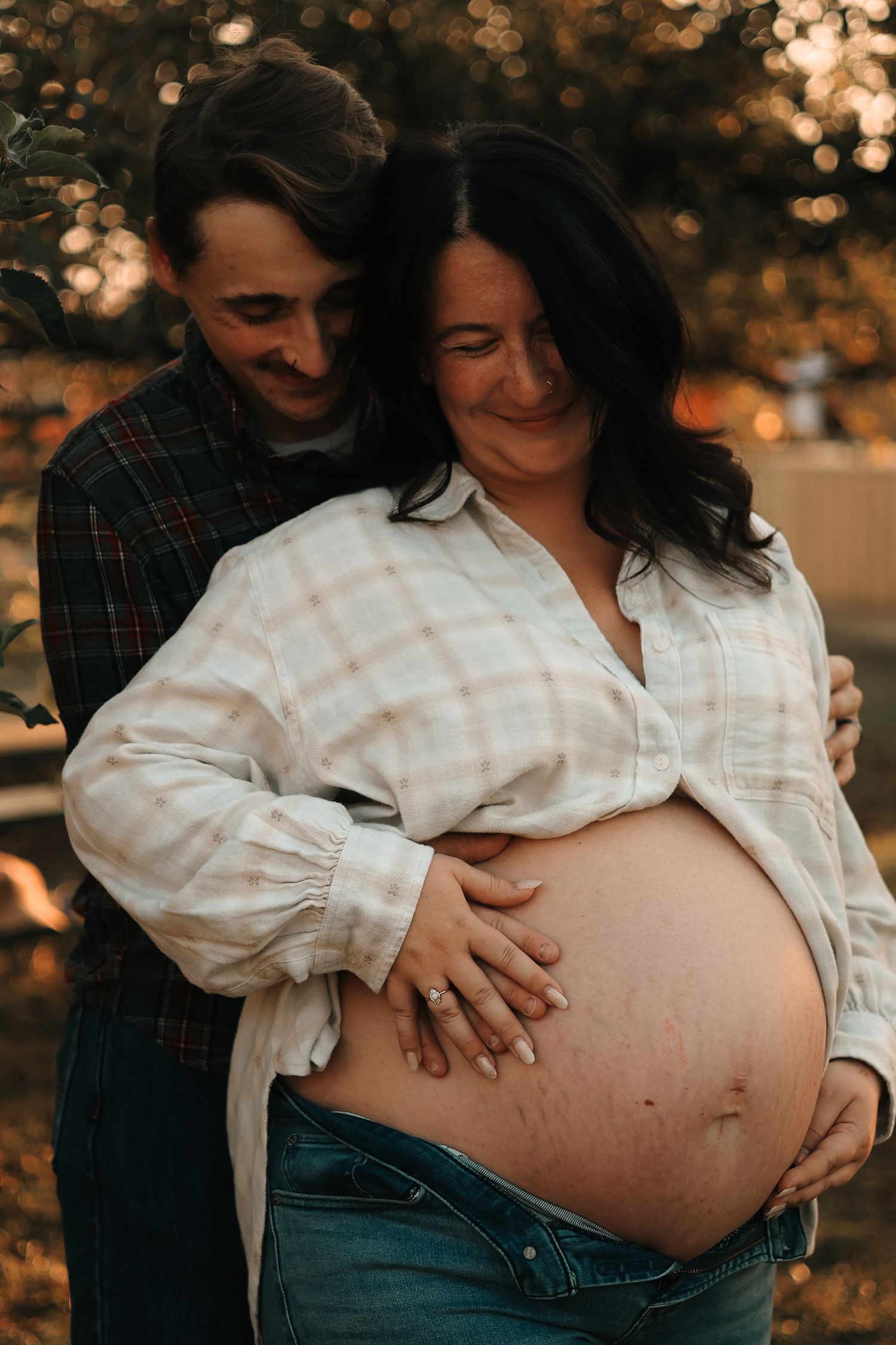 Couple embracing at sunset, man nuzzling pregnant partners neck while both cradle her large baby bump tenderly.