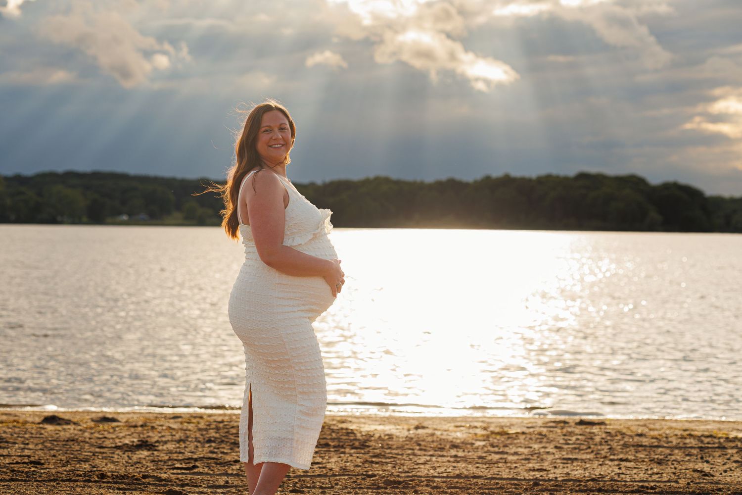 Pregnant woman in white dress standing on beach at golden sunset with rays of light over water.