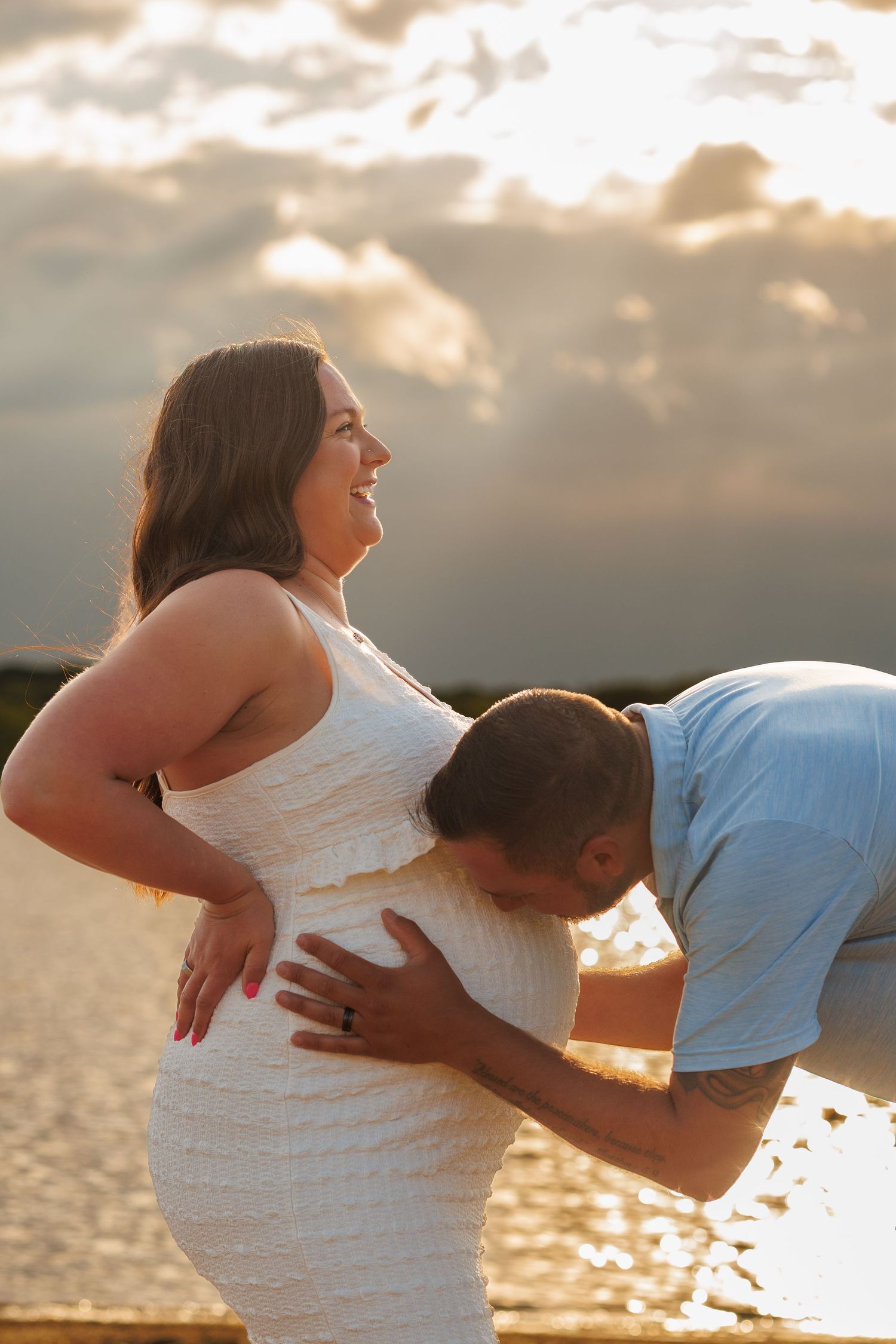 A man kisses a pregnant woman's belly outdoors at sunset near water, as she smiles joyfully in a white dress.