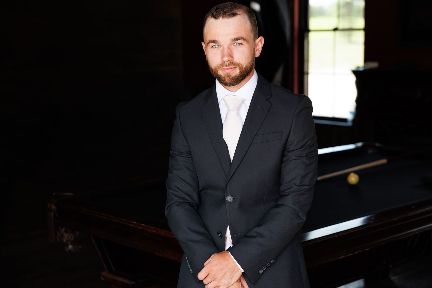 Man in black suit with white tie standing confidently in front of a pool table in a dark elegant room.