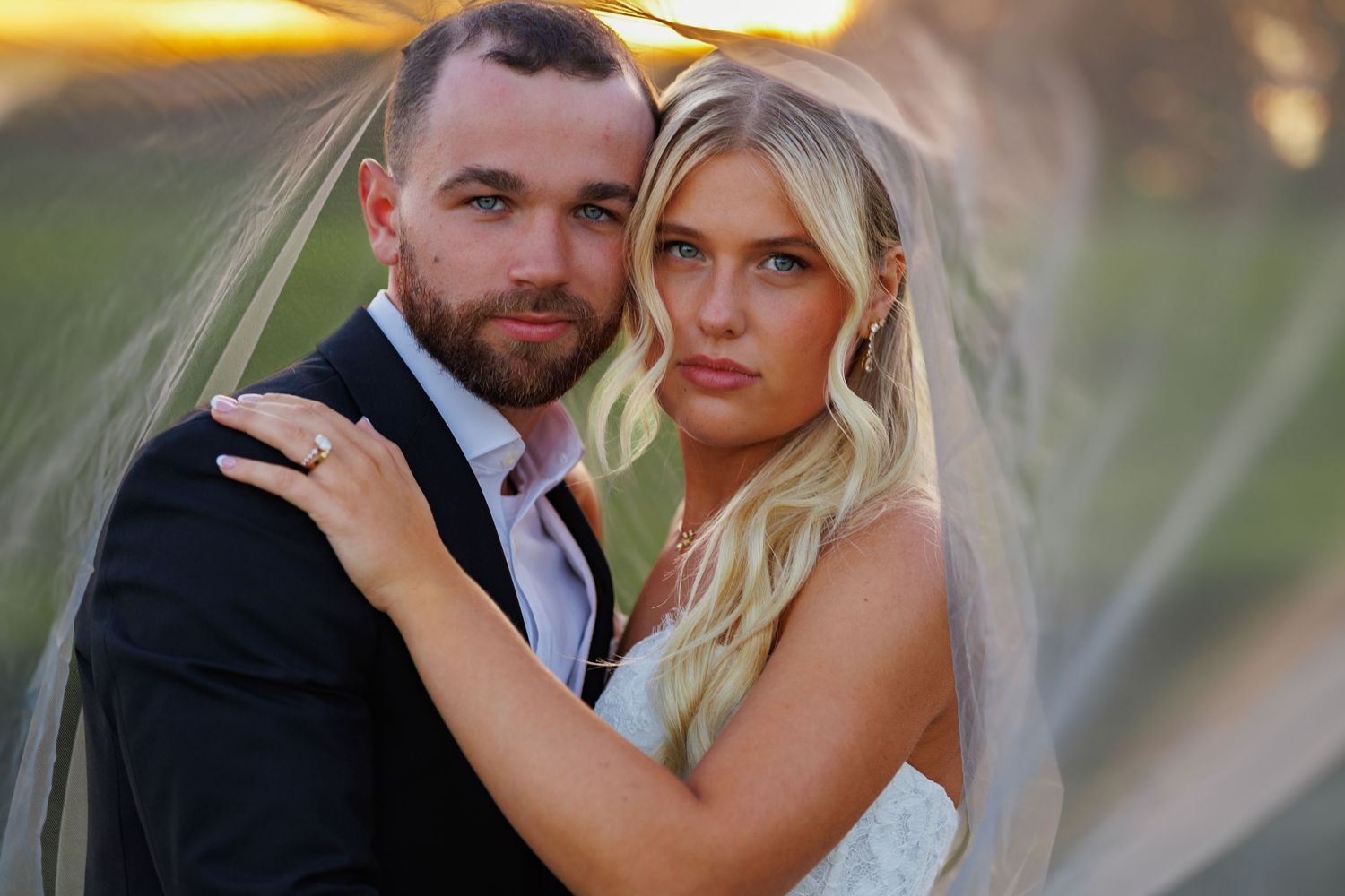 A bride and groom pose romantically under a flowing veil at golden hour during their outdoor wedding.