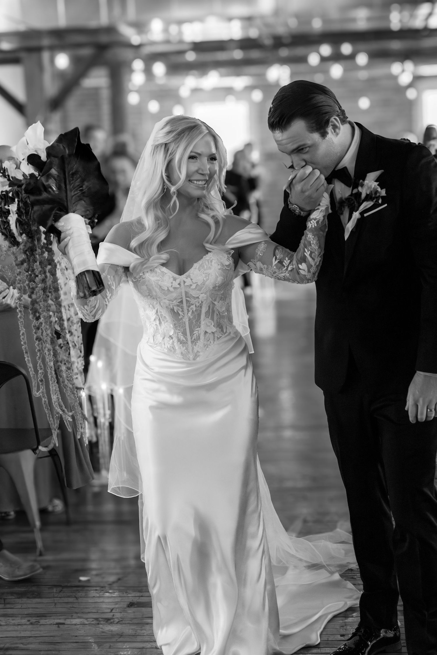 Groom kissing bride's hand as they walk down the aisle in a black and white wedding photo.