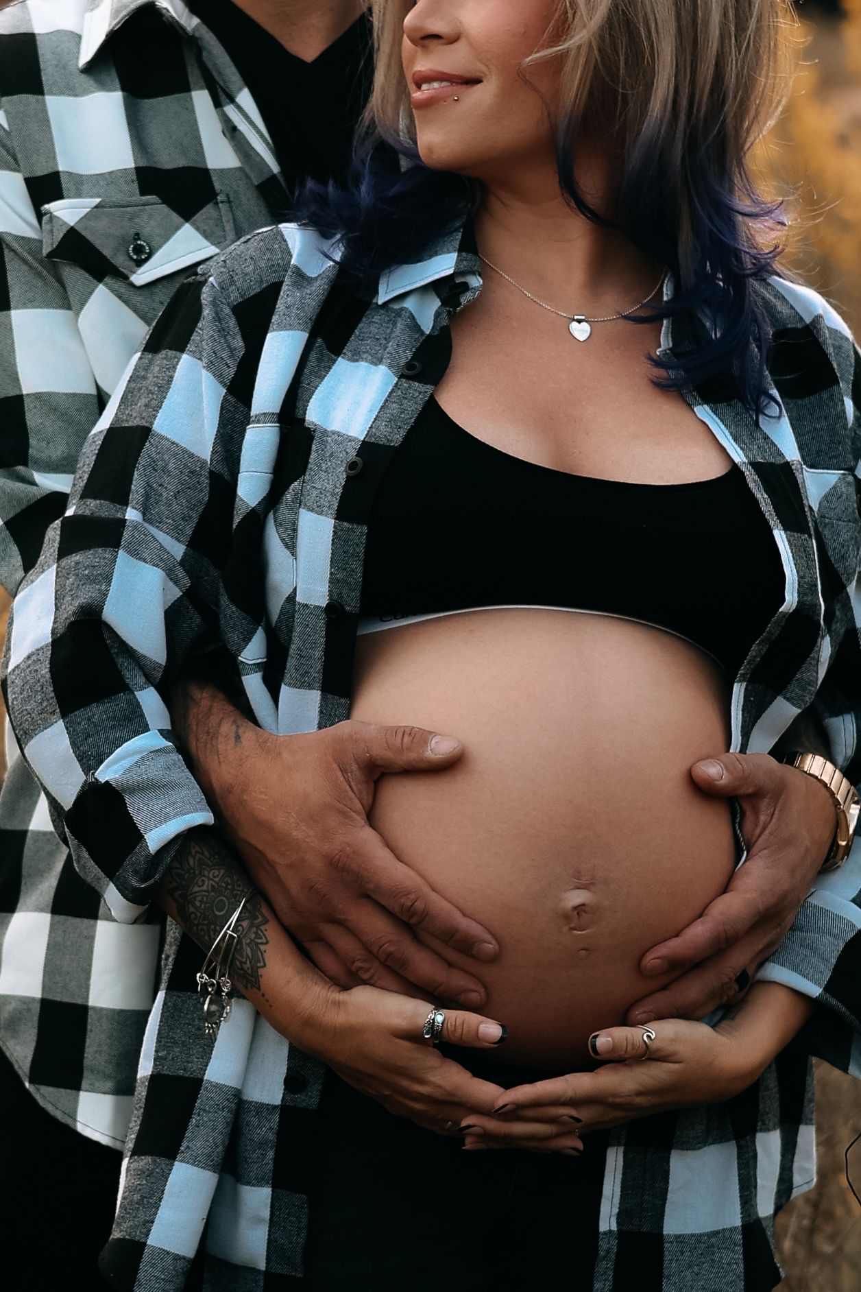 Close-up of couple in plaid shirts cradling pregnant belly with both hands outdoors.