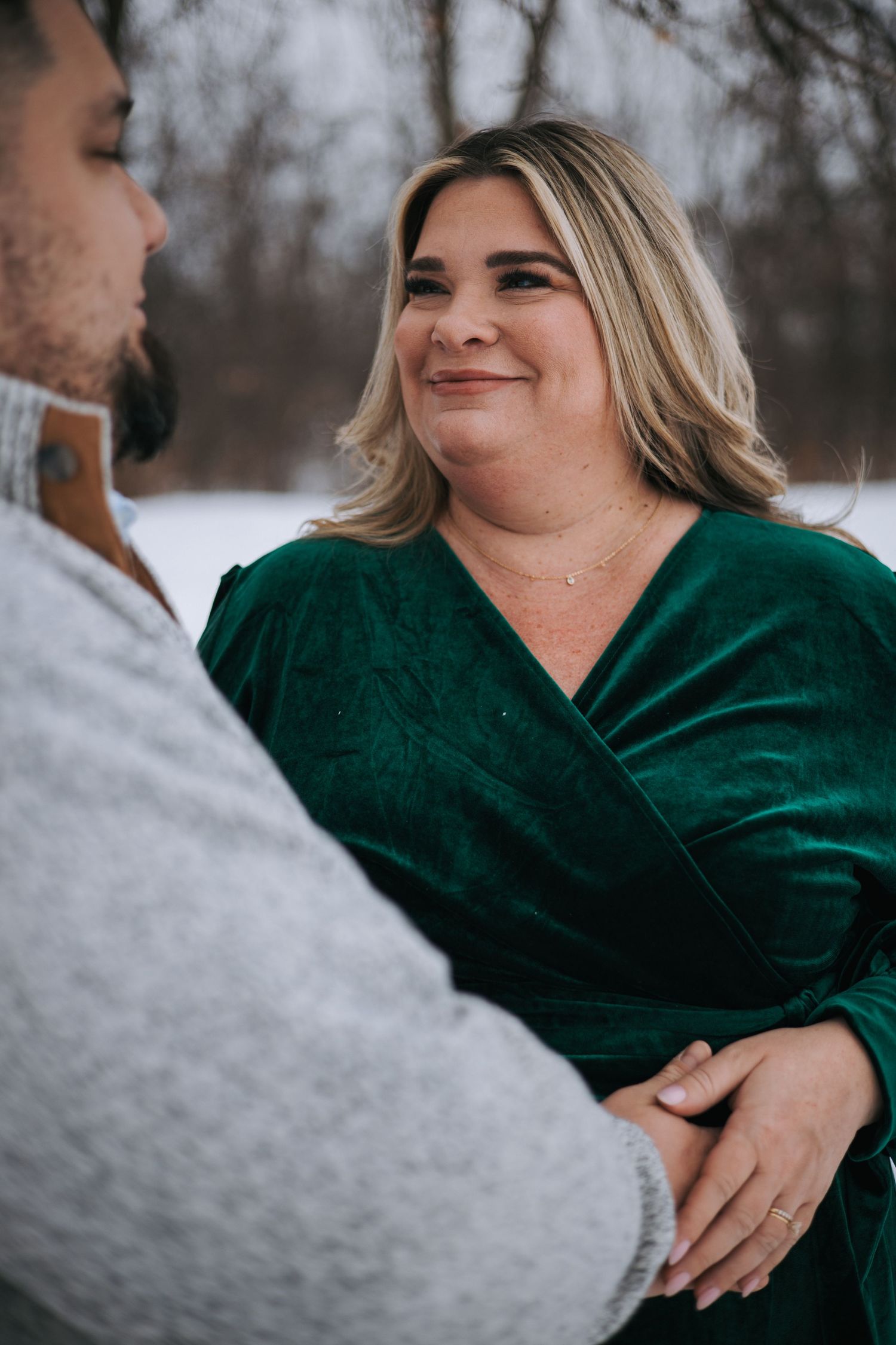 Pregnant woman in green velvet dress smiling warmly at partner while holding hands in winter snow.