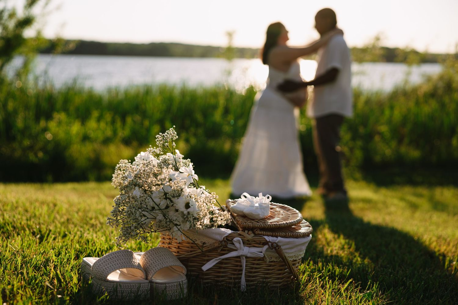 Couple dancing by lakeside at sunset with white floral bouquet and wicker basket in foreground on green grass.