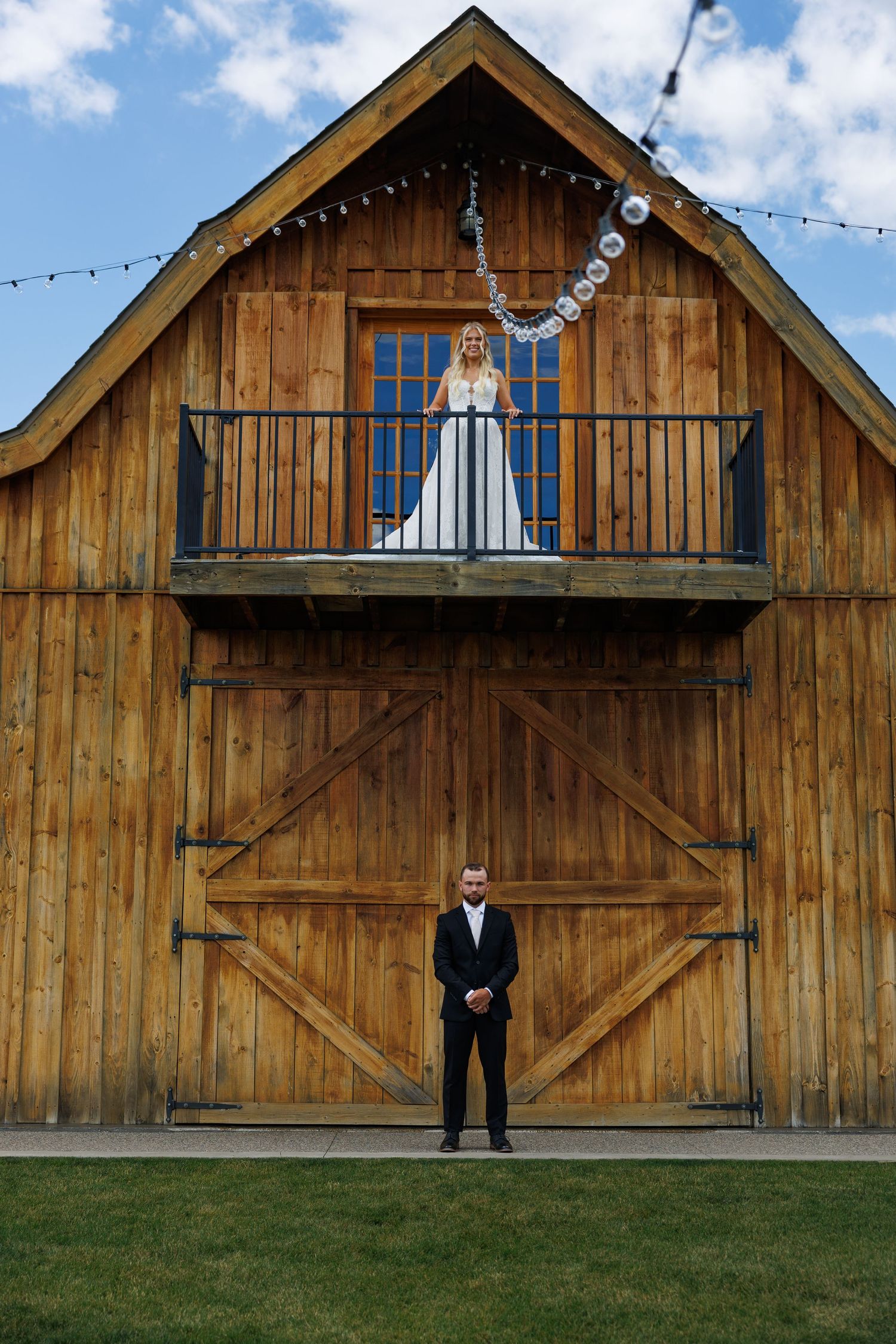 Bride on barn balcony with groom standing below at rustic wooden barn wedding venue under blue sky.