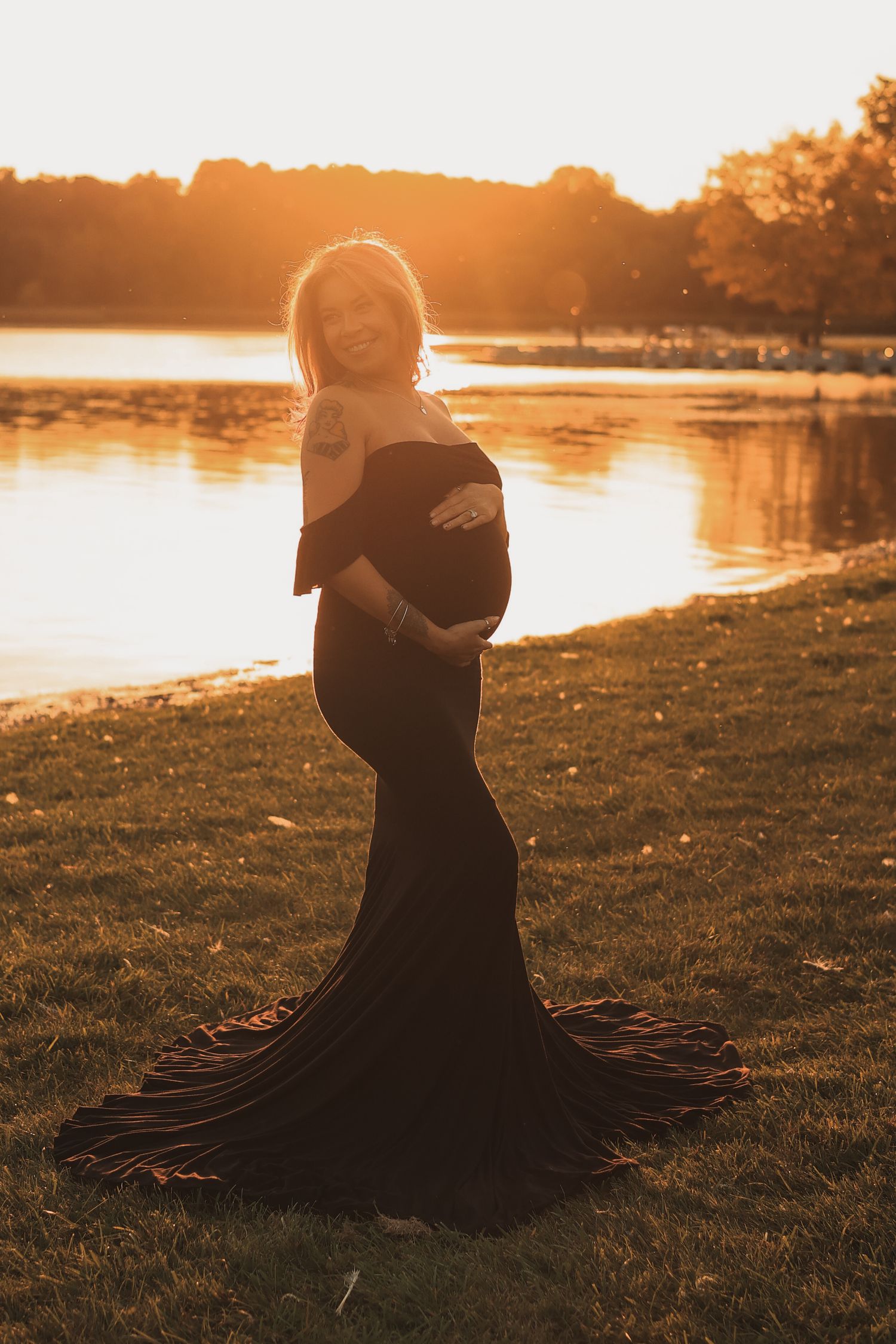 Pregnant woman in elegant black off-shoulder gown posing by a lake during a glowing sunset.
