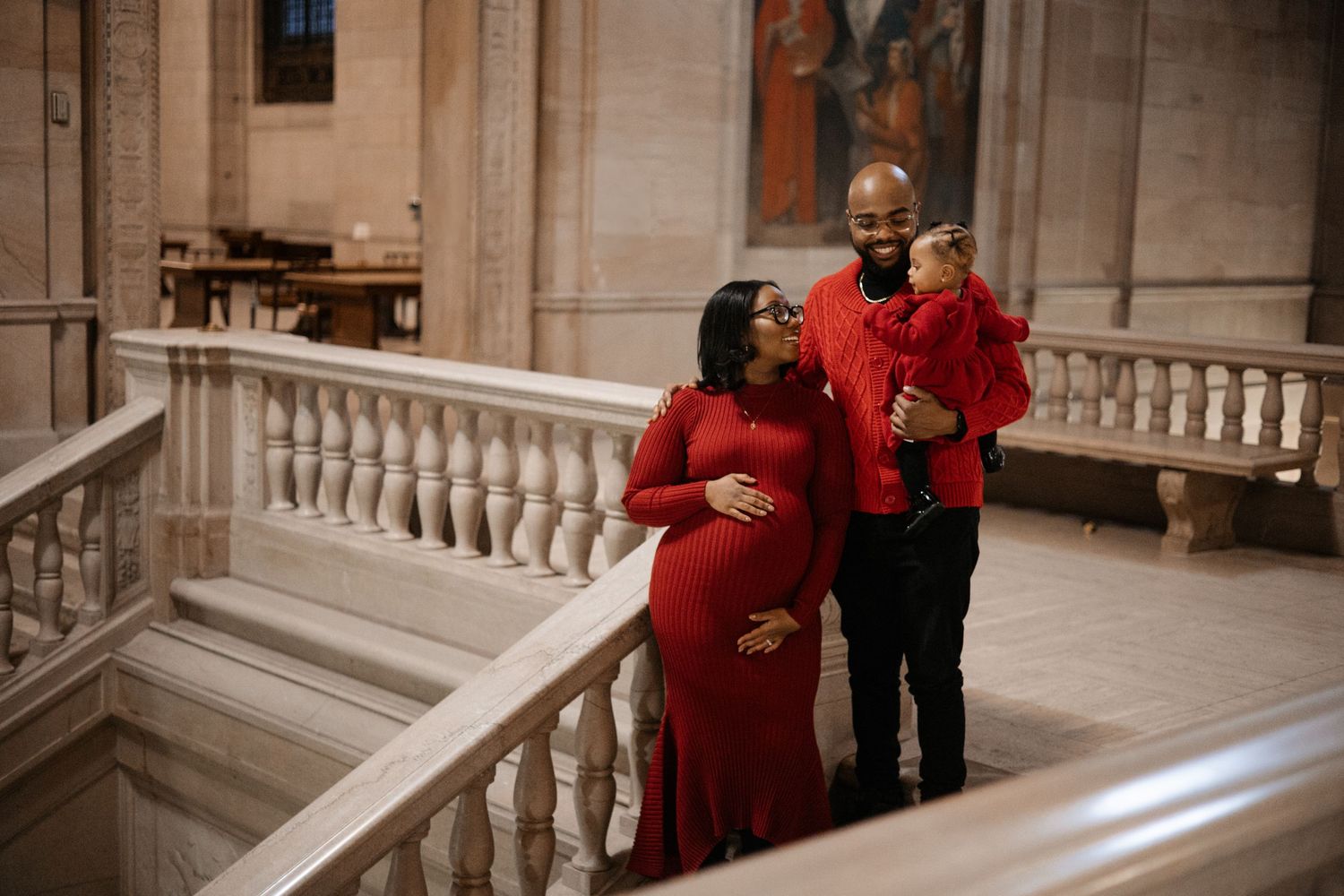 A pregnant woman in a red dress poses with her partner and toddler on marble stairs in an elegant building.