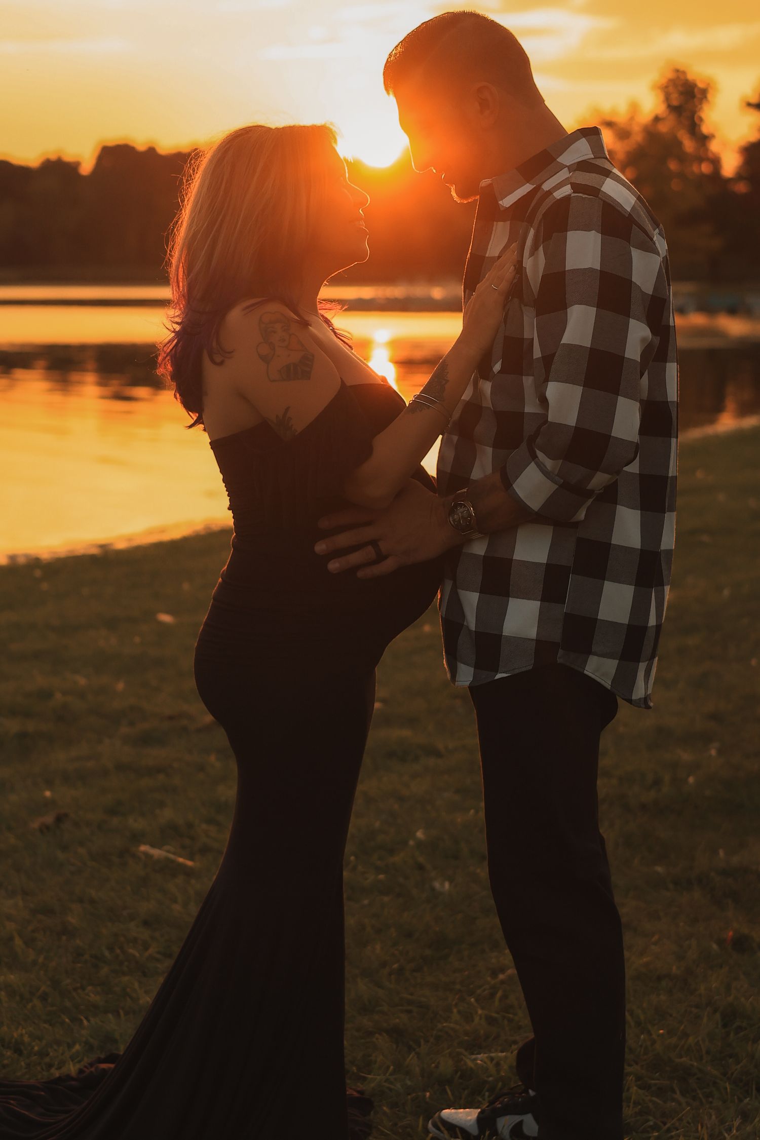 Silhouetted pregnant couple facing each other intimately by a reflective lake during a stunning orange sunset.