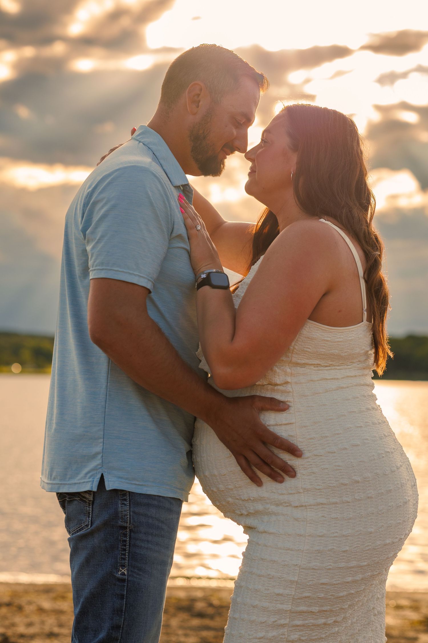 Pregnant couple sharing a tender moment by a lake at golden sunset, woman in white dress with partner in blue shirt.