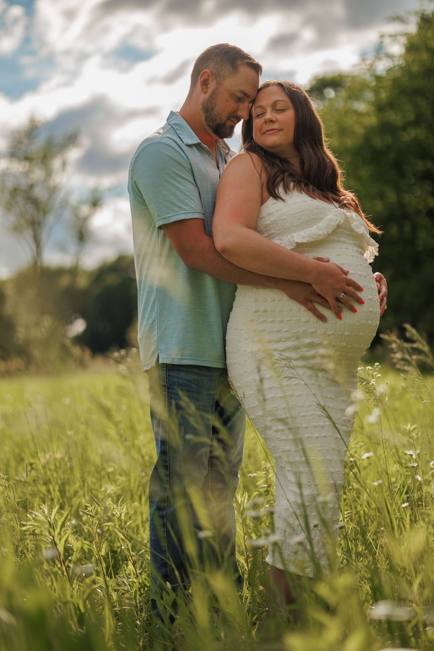 Couple embracing in a sunny green field, pregnant woman in white dress with partner in blue polo.
