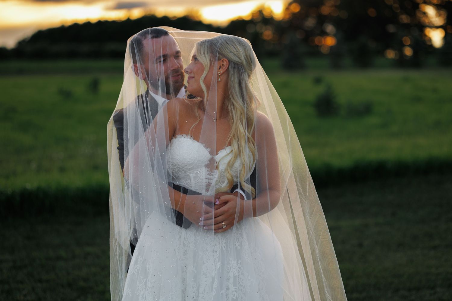 Bride and groom share romantic moment under wedding veil at sunset on a lush green countryside backdrop.