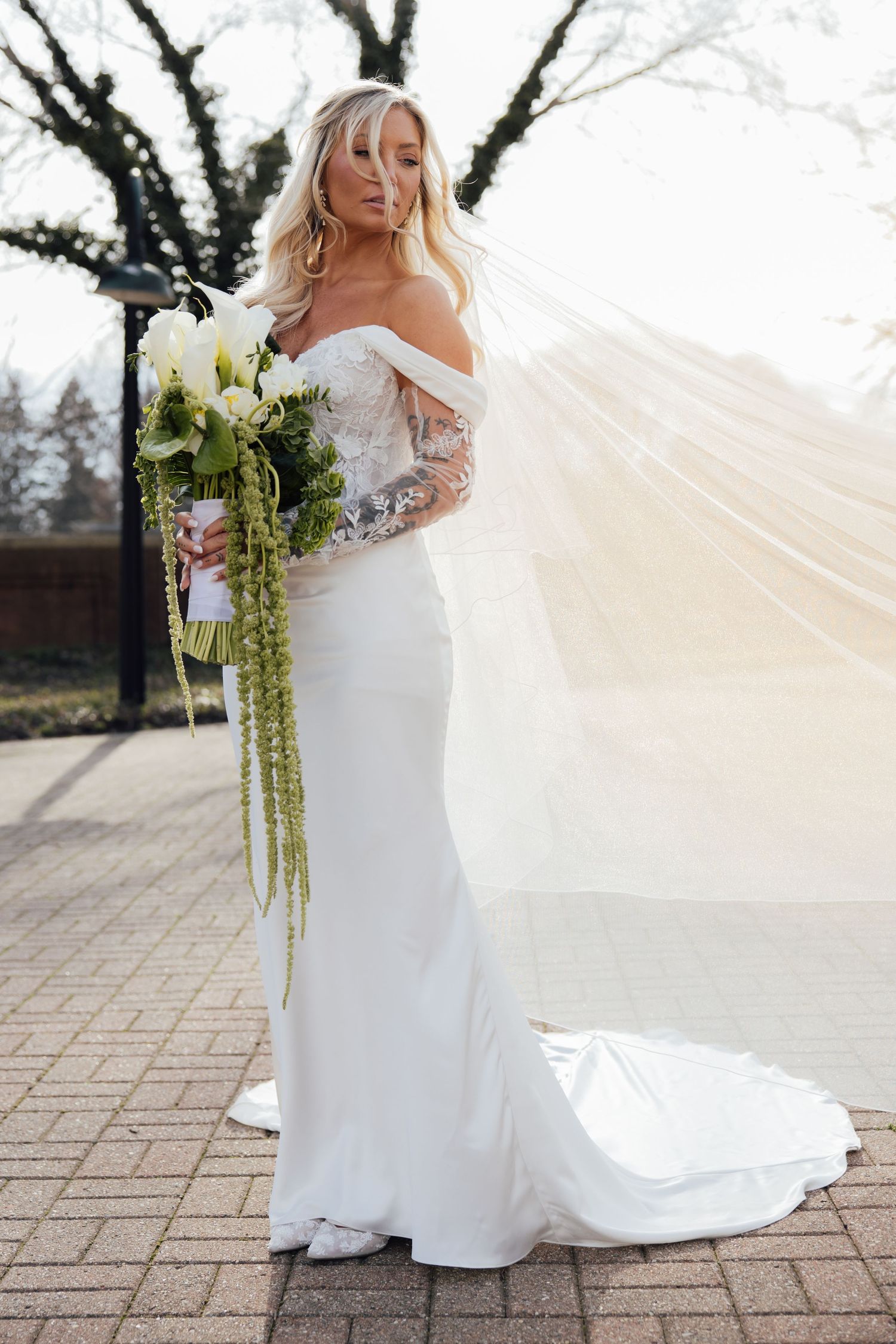 Bride in off-shoulder lace mermaid gown holding cascading green and white bouquet with veil billowing in wind.