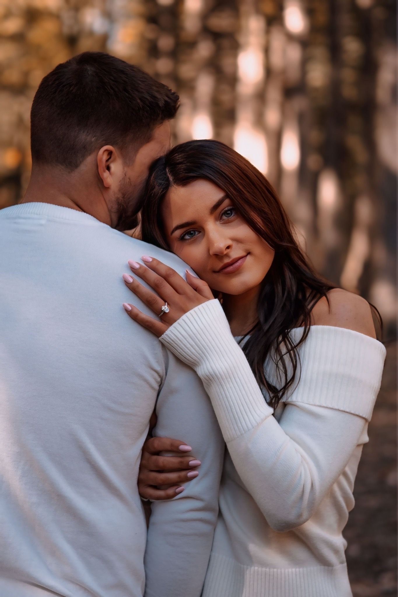 Couple embracing in autumn forest, woman in white off-shoulder sweater showing engagement ring while resting head on mans chest.