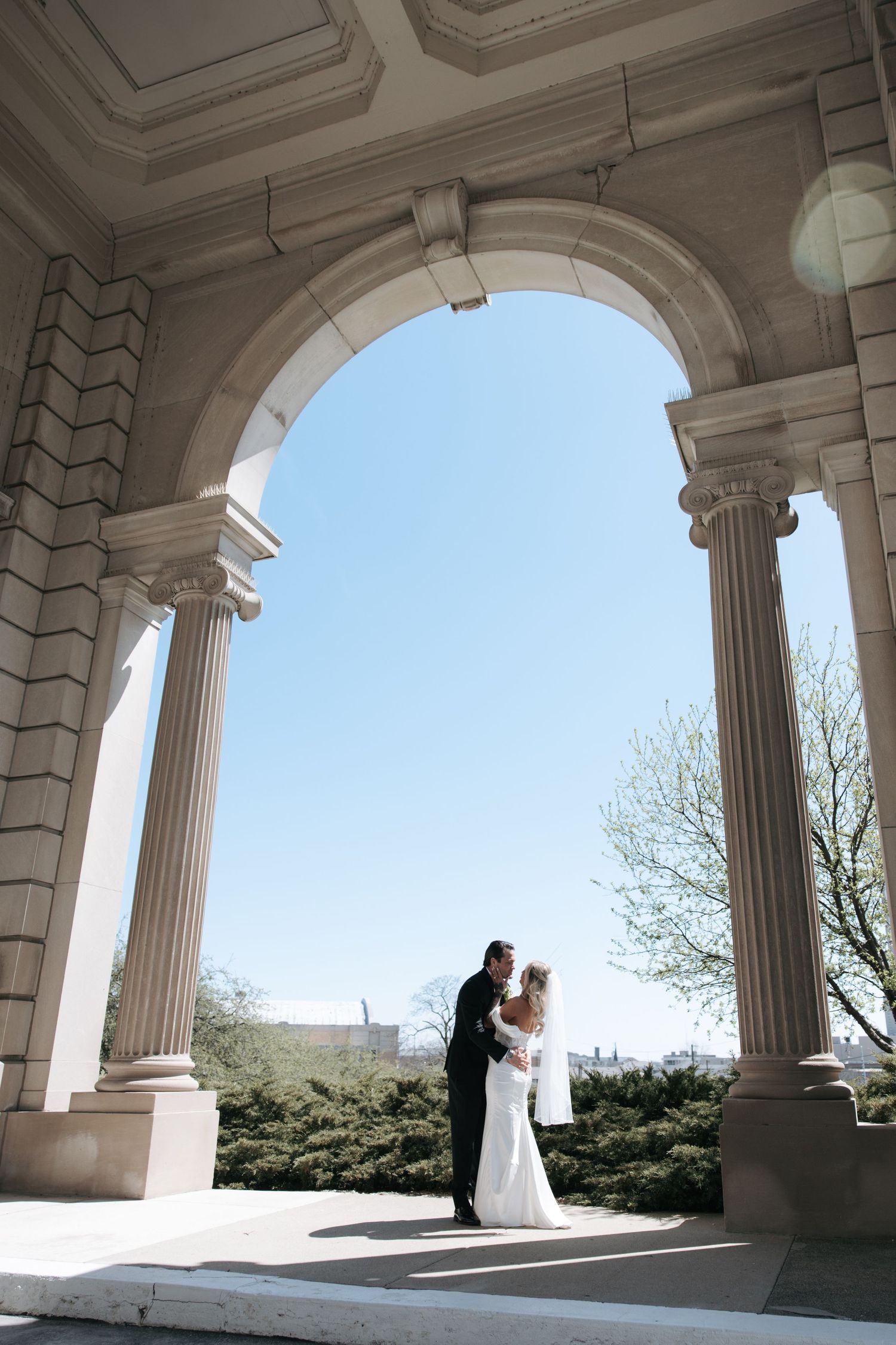 Bride and groom share a romantic moment under elegant stone arches with columns at a classic wedding venue.