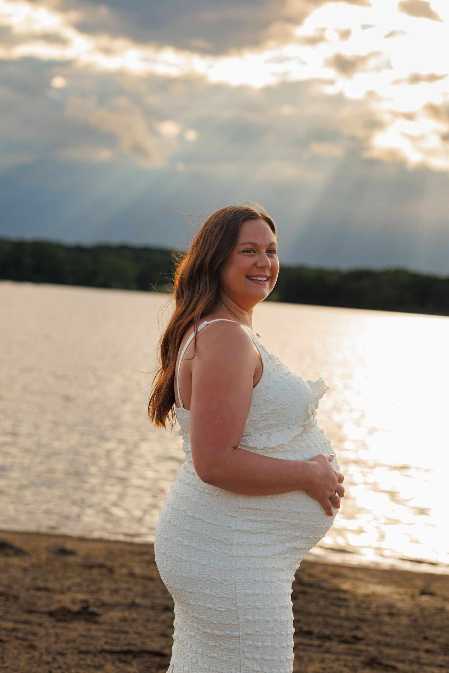 Smiling pregnant woman in white dress holding belly on beach at golden sunset with dramatic cloudy sky.