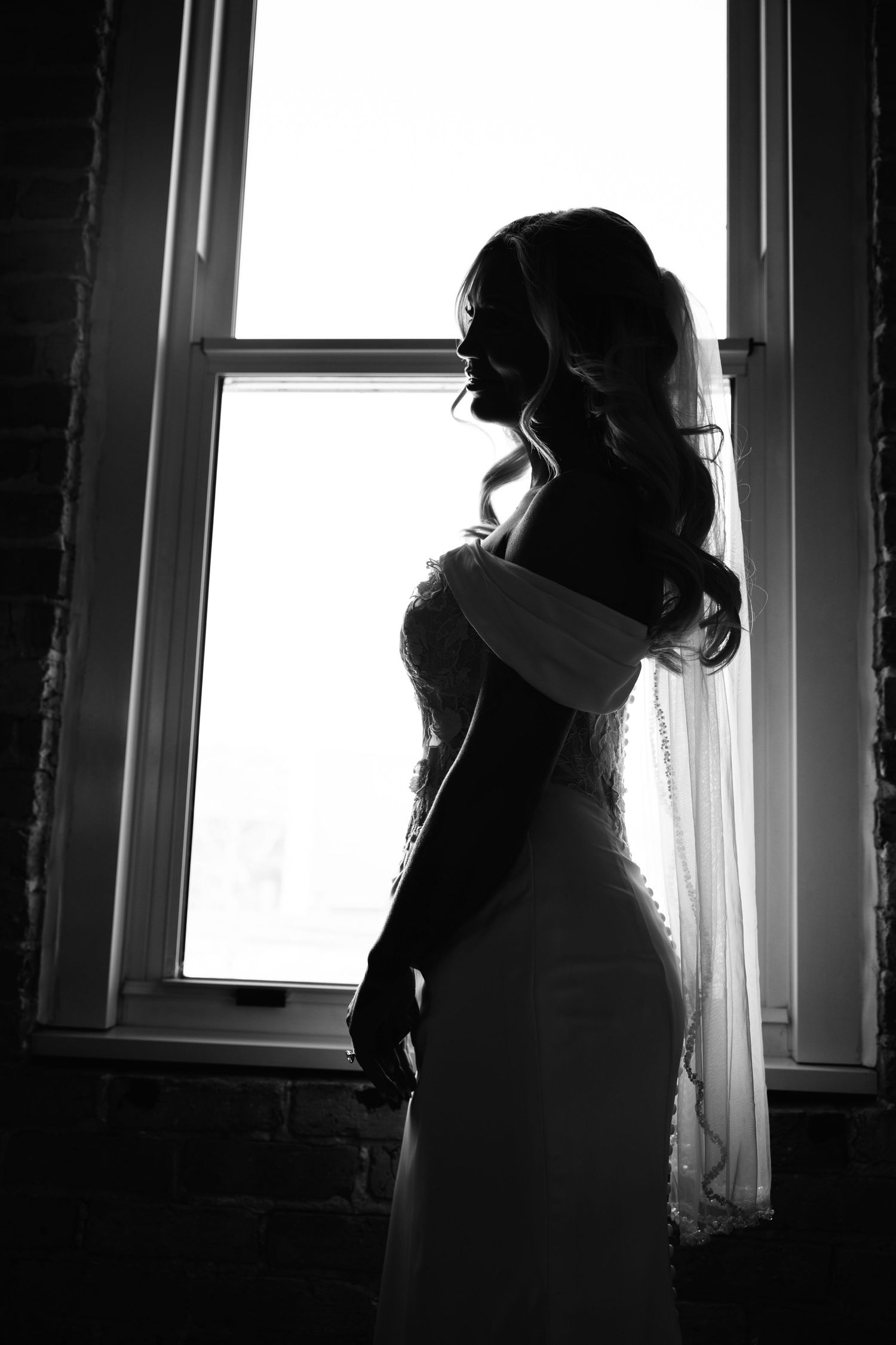 Elegant bride in wedding gown and veil silhouetted against a bright window in black and white.