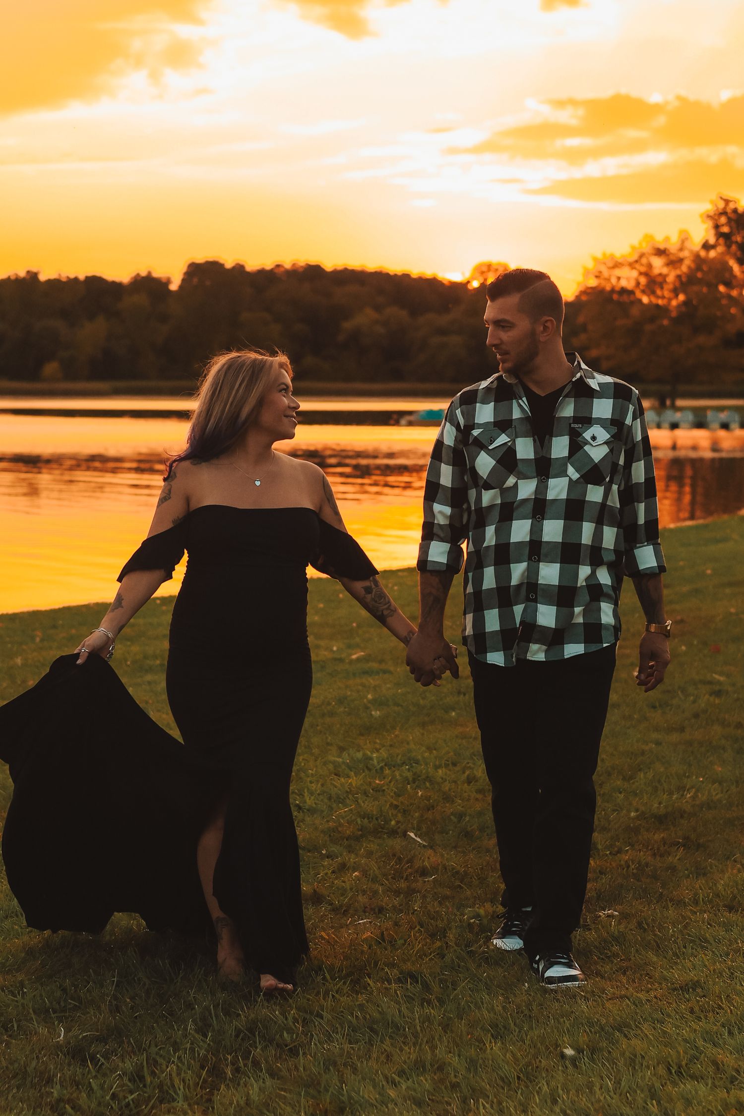 Pregnant couple holding hands and walking by a lake at vibrant golden sunset, woman in flowing black dress.