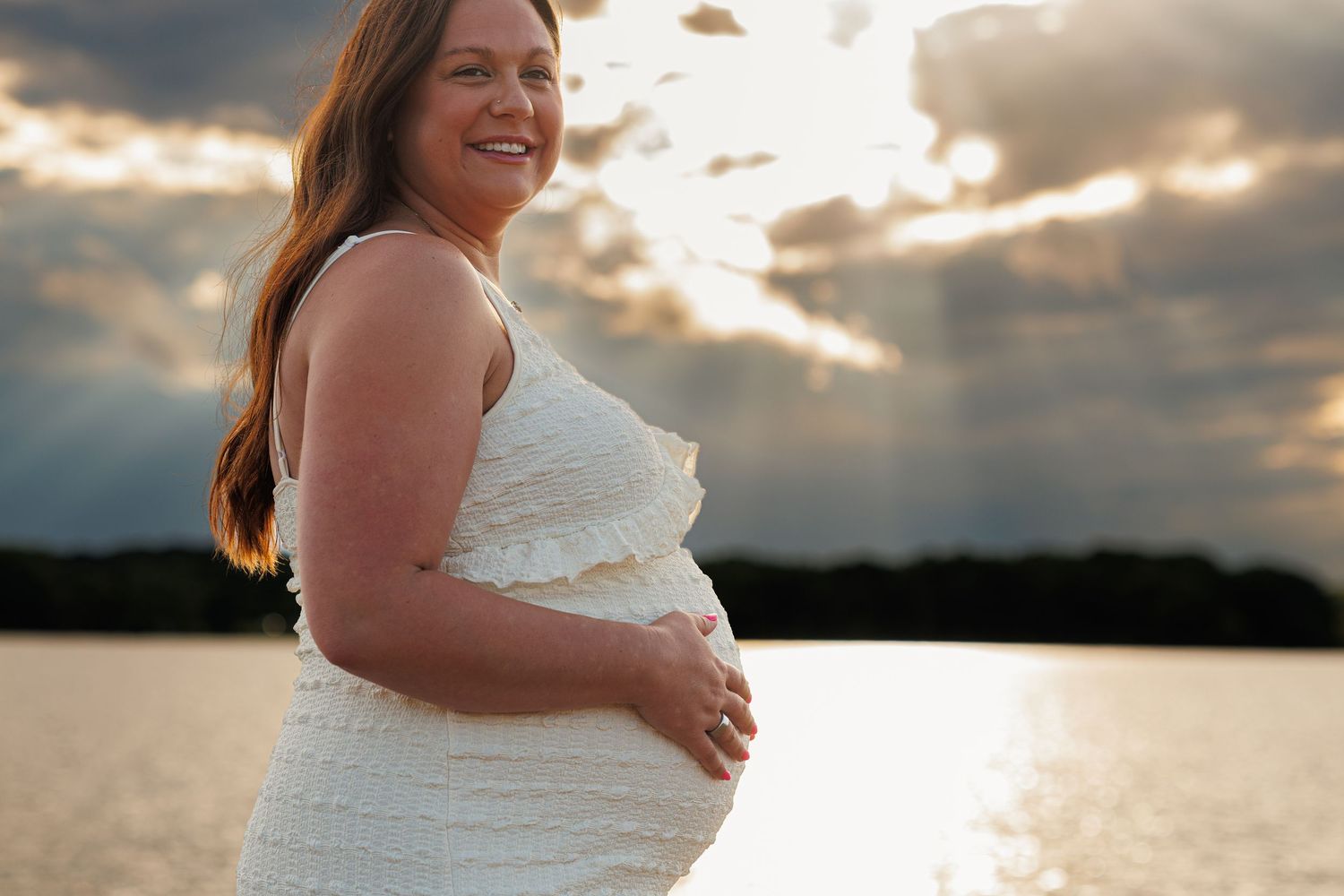 Smiling pregnant woman in white dress posing by a lake at sunset with dramatic cloudy sky.