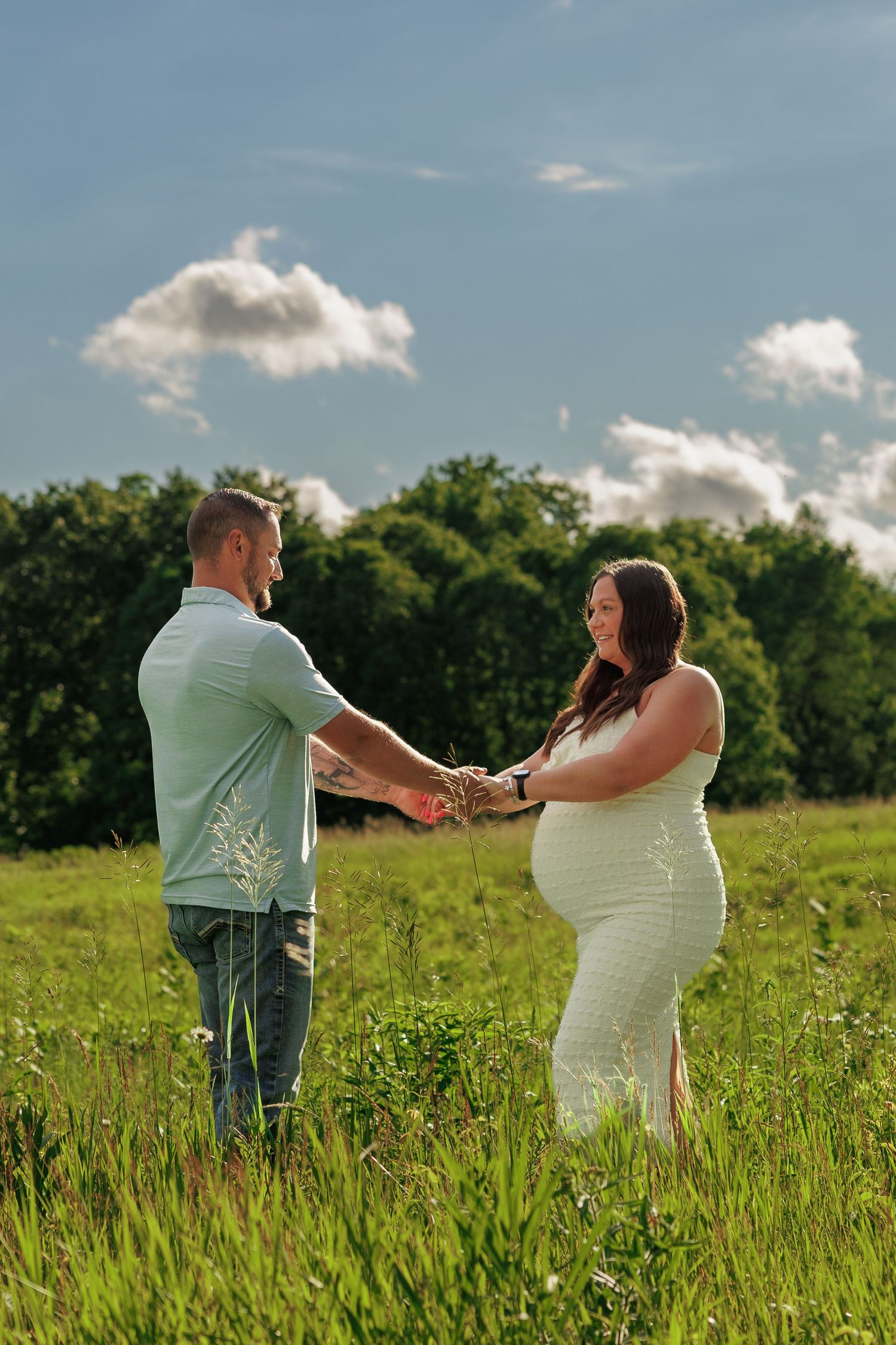 Pregnant couple holding hands in a green field under a blue sky, sharing a tender maternity photo moment outdoors.