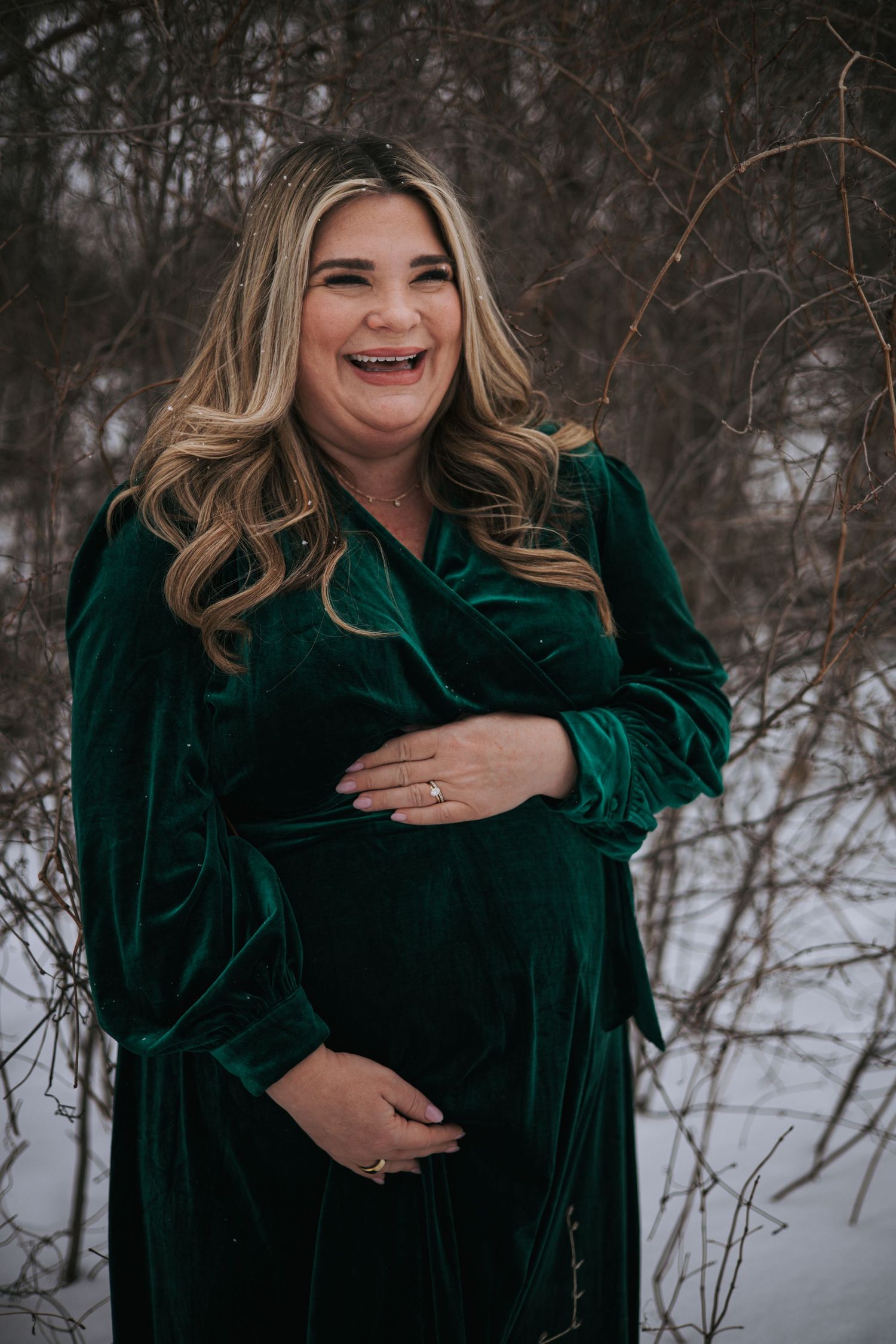 Smiling pregnant woman in emerald green velvet dress holding baby bump outdoors in winter snow.