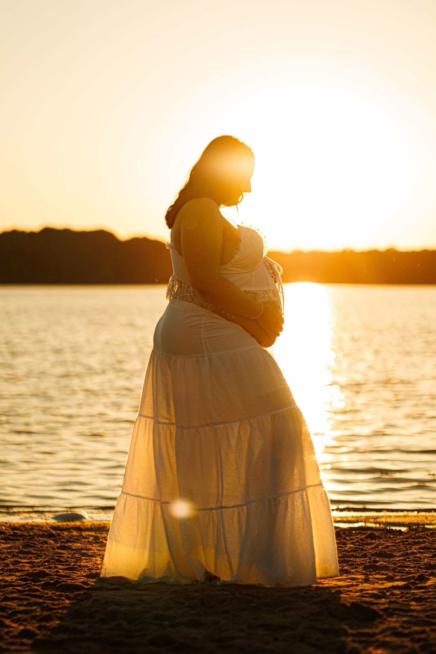 Pregnant woman in white dress standing on beach at golden sunset, cradling baby bump by serene lake.