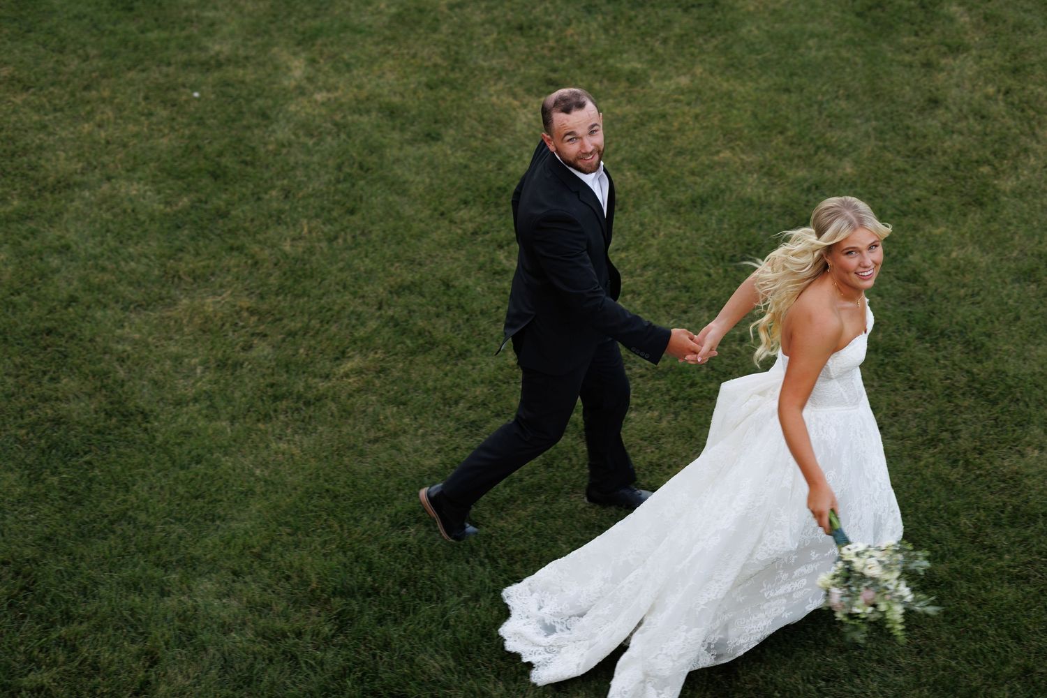 Aerial view of bride in white gown and groom in black suit holding hands while walking on green grass.